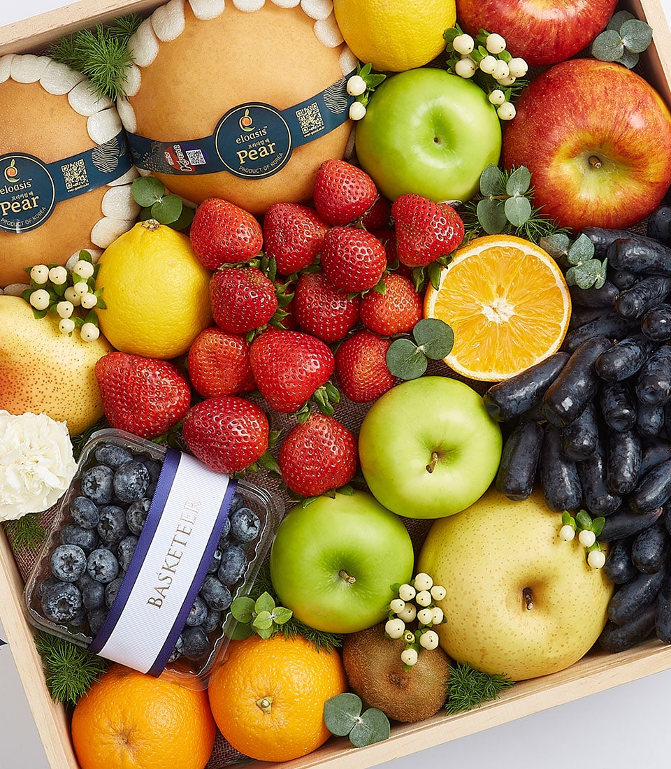 A luxurious wooden crate brimming with an exquisite selection of farm-fresh fruits, carefully curated by Basketeer. Among the bounty, you'll find two premium Korean pears, crisp green apples, succulent red apples, vibrant strawberries, plump blueberries, luscious grapes, and juicy oranges—including a perfectly half-sliced orange. This opulent arrangement is further adorned with delicate white flowers and lush green leaves creating a visually stunning gift of fruit elegance. A tasteful ribbon featuring the Basketeer logo adds an extra touch of sophistication to this gourmet treat.