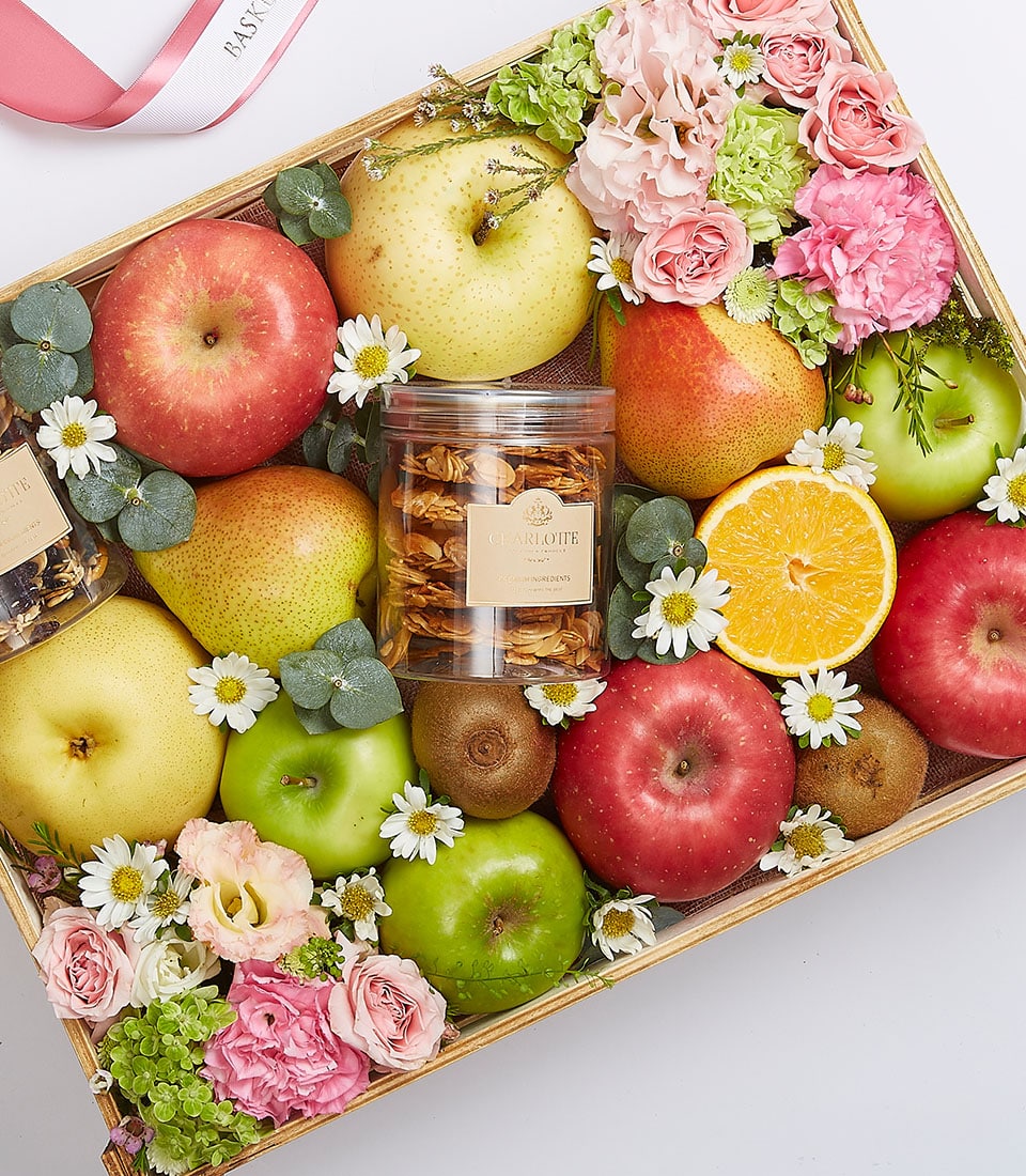 A lavish wooden tray laden with a sumptuous collection of apples, pears, kiwis, and a vibrant orange half. Complemented by an elegant jar of dried fruit and encircled by delicate pink and white flowers along with lush greenery. The scene is crowned by a glimpse of the luxurious Basketeer ribbon at the top left corner—an authentic Exotic Fruit Gift Box delight that epitomizes sophistication and elegance.