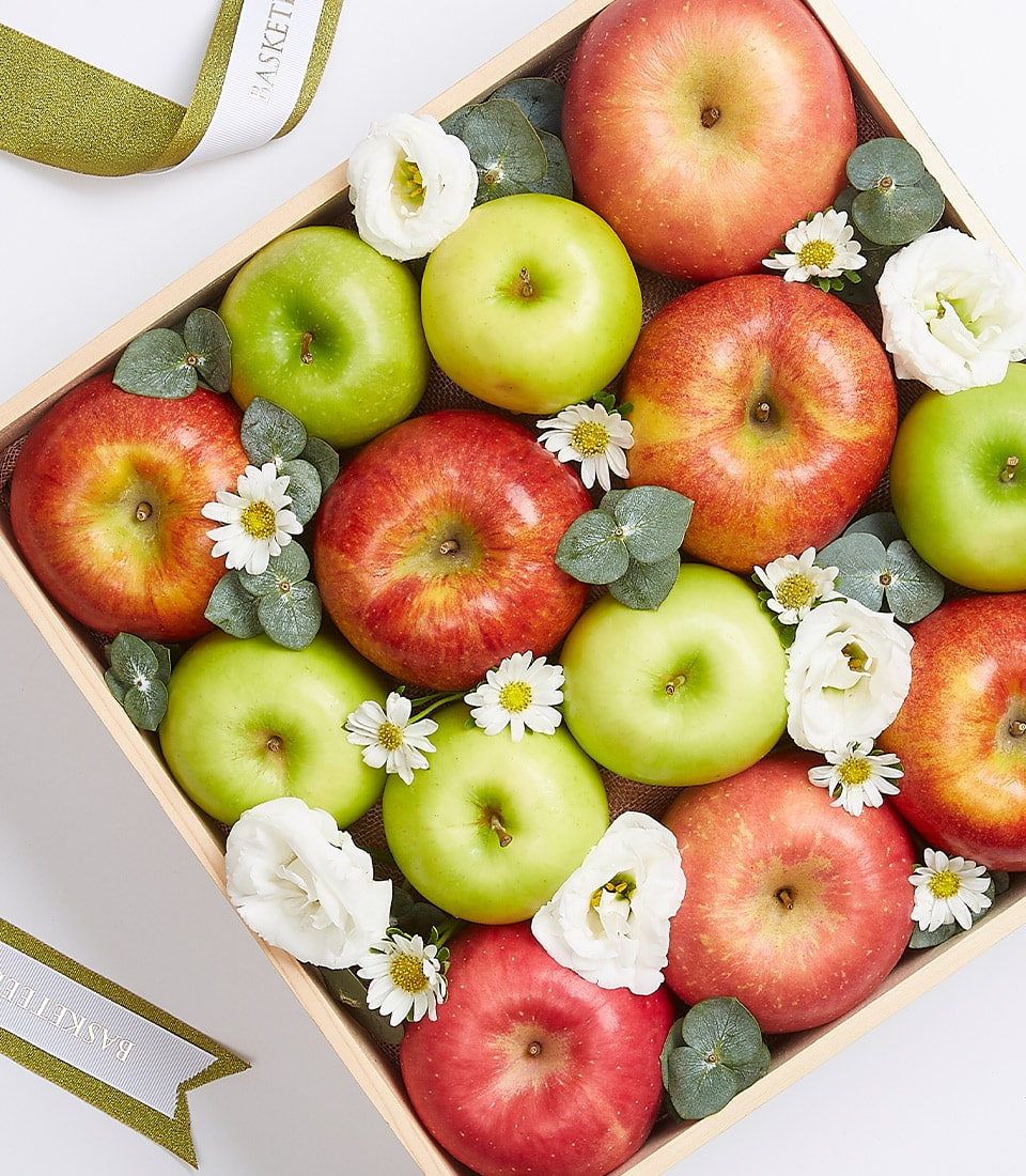 An opulent wooden Apple Gift Box Royale, brimming with a luscious assortment of vibrant red and green apples, complemented by delicate white flowers and lush green leaves. This exquisite arrangement is captured from a top-down view, inviting admiration. A luxurious ribbon adorned with 