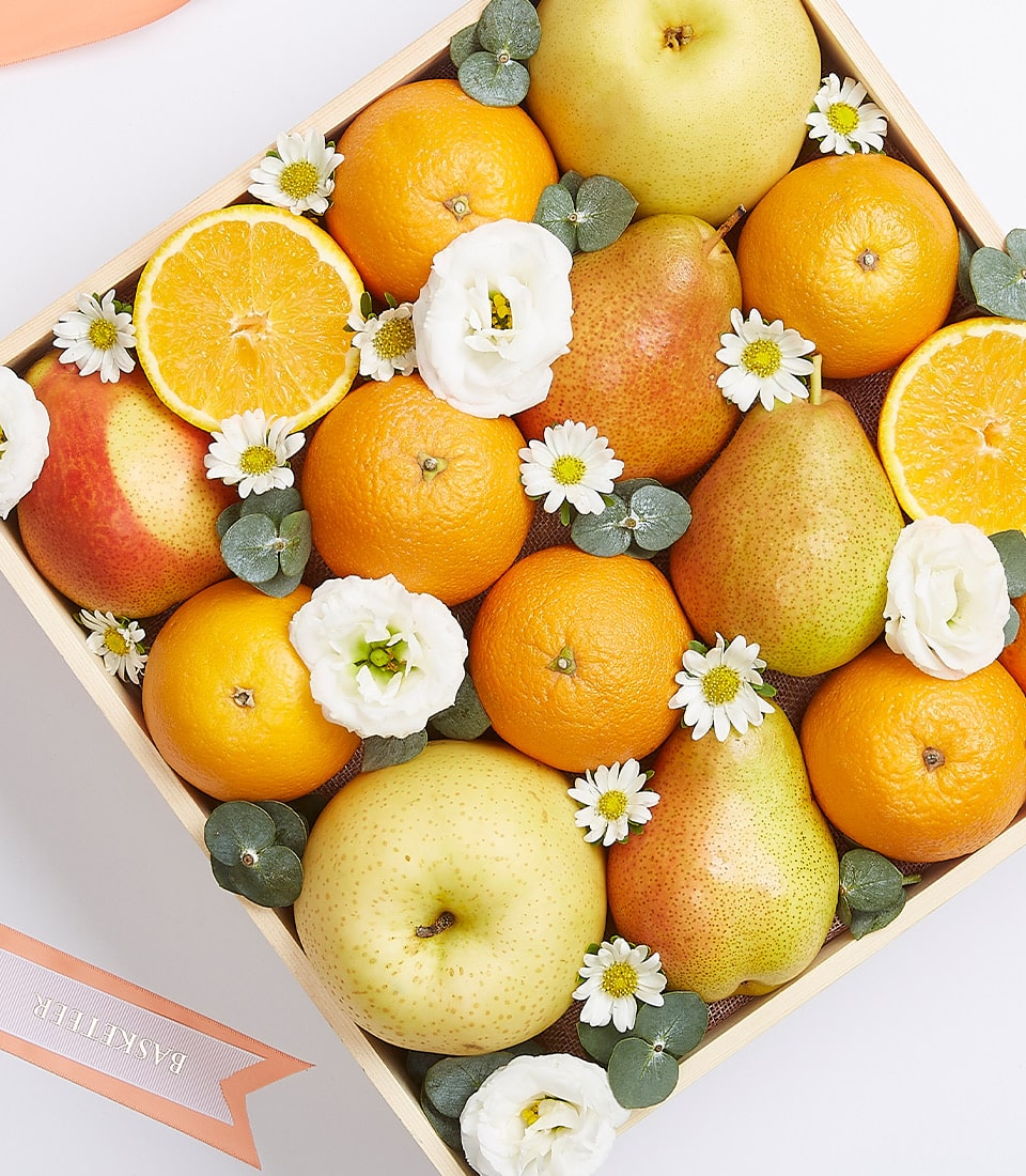A luxurious Basketeer fruit gift box exudes elegance and sophistication. Inside, a curated selection of the freshest oranges, apples, and pears is beautifully arranged. Each piece of fruit nestles among delicate white flowers and lush green leaves. The crate is adorned with a sumptuous peach-colored ribbon that cinches the scene perfectly. One orange is artfully cut in half, revealing its luscious, juicy segments—a true testament to the quality within. The iconic Basketeer logo graces the ribbon and box, adding a touch of exclusivity to this already inviting presentation.