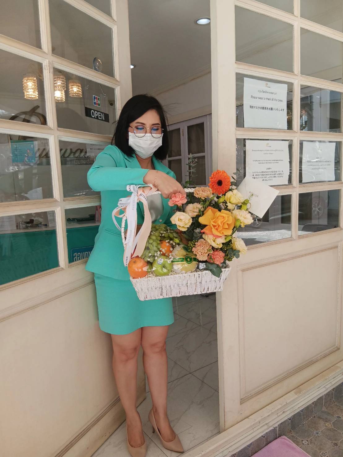 A woman wearing a green outfit and face mask stands in an open doorway, holding a white basket filled with various colorful flowers, including yellow blooms and ribbons. A sign with text is visible on the glass door behind her.
