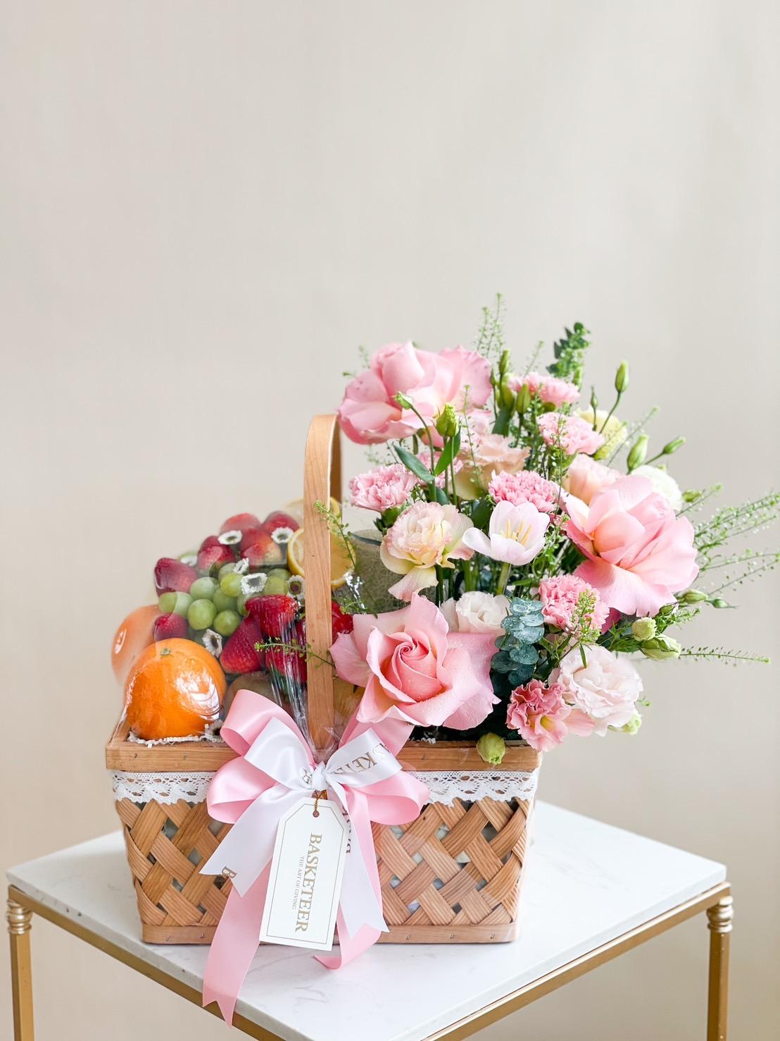 A wicker gift basket with a handle sits on a white table. It contains mixed fruits including oranges, grapes, and strawberries on one side, and a vibrant arrangement of pink and white flowers on the other. A pink ribbon with a tag reading 