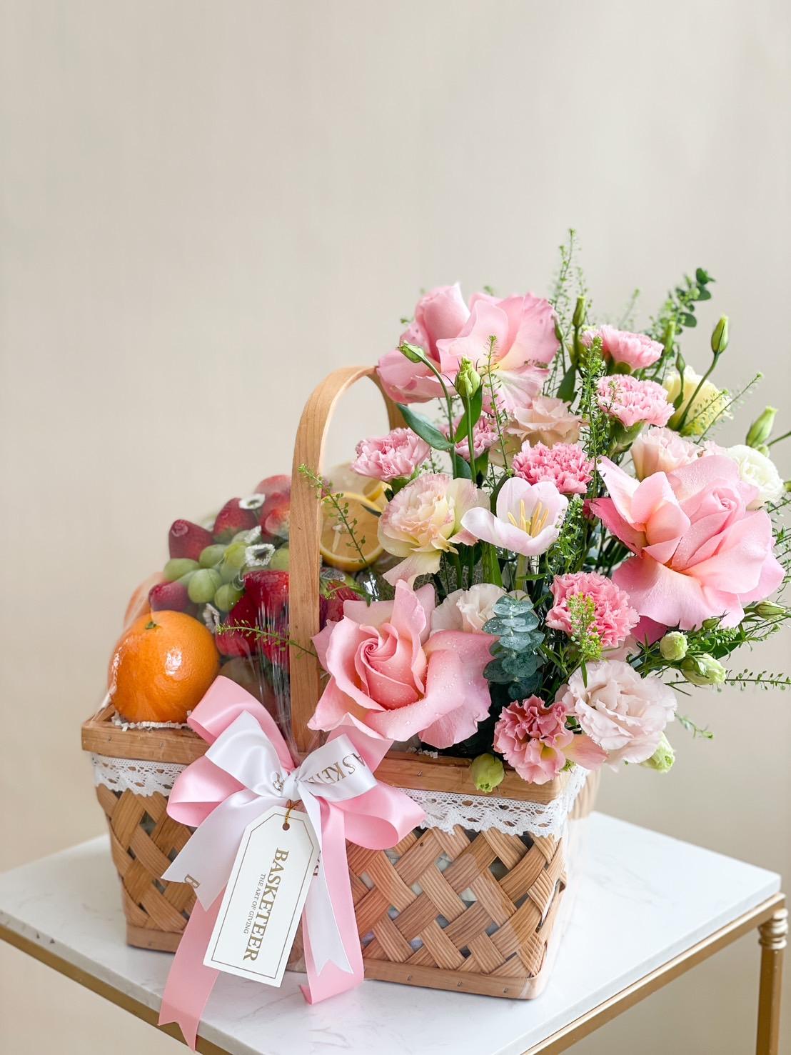 A wicker basket containing a colorful arrangement of pink roses, carnations, and mixed flowers, as well as a variety of fresh fruits like oranges, green grapes, and red grapes. The basket is embellished with a white bow and ribbon, with a 
