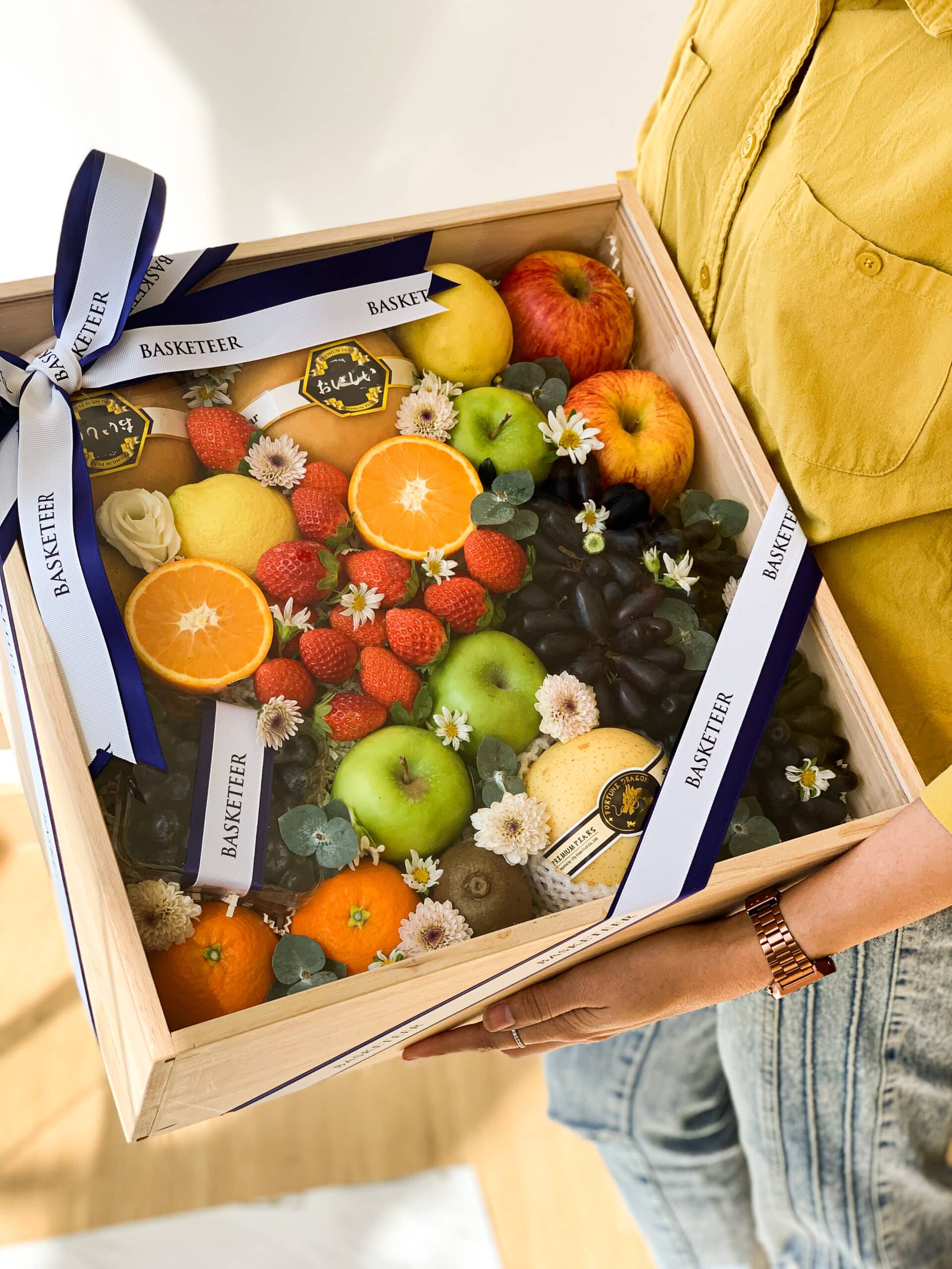 A person wearing a yellow shirt and jeans holds a bountiful Fruit Elegance gift box adorned with a blue and white ribbon. The wooden box contains an assortment of fresh fruits such as apples, grapes, oranges, strawberries, green apples, and decorative flowers.