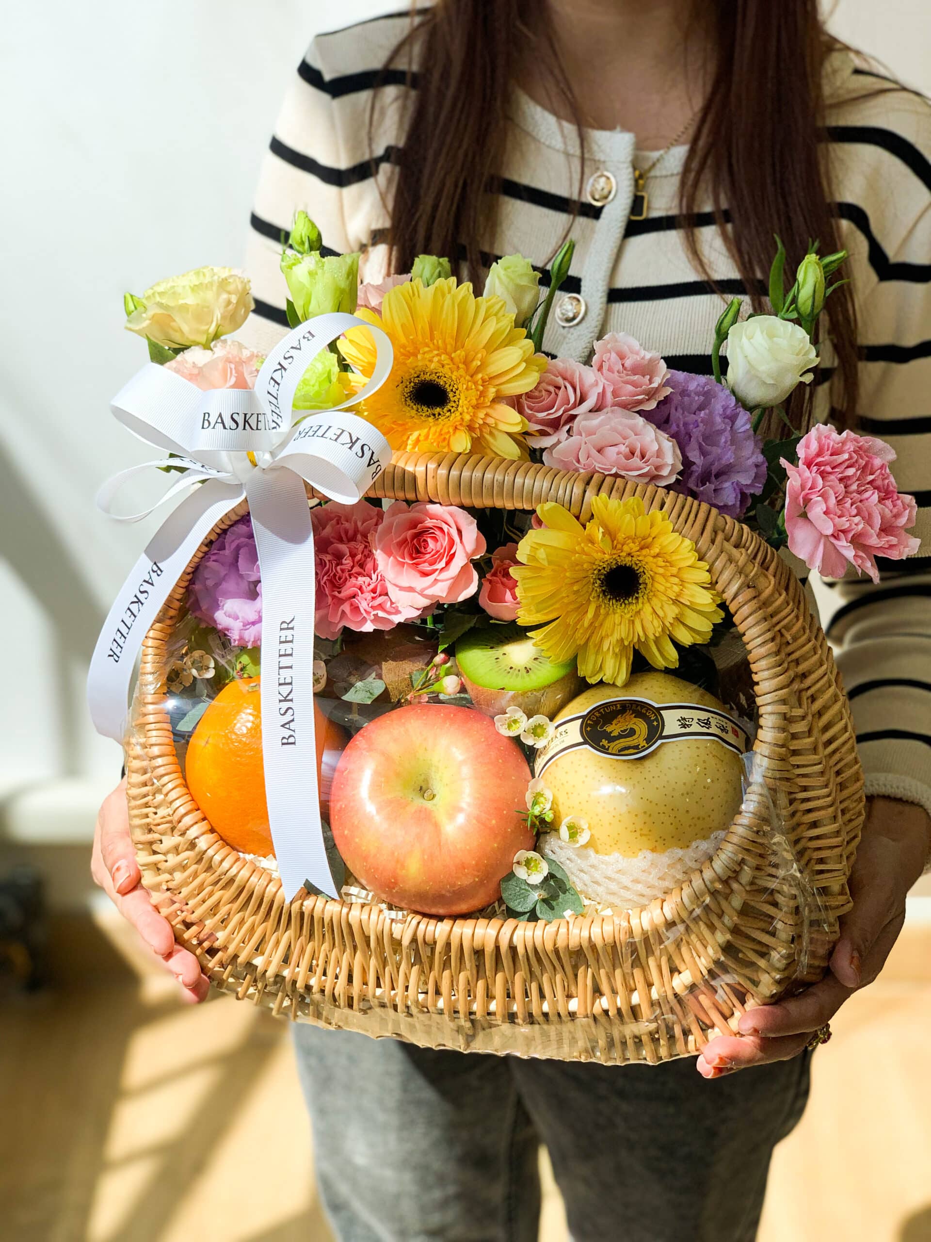 A person holding a Luscious Fruit Harvest Basket adorned with yellow daisies, pink roses, and purple carnations. The basket also contains a selection of fruits such as apples, pears, and oranges. The person is wearing a striped sweater.