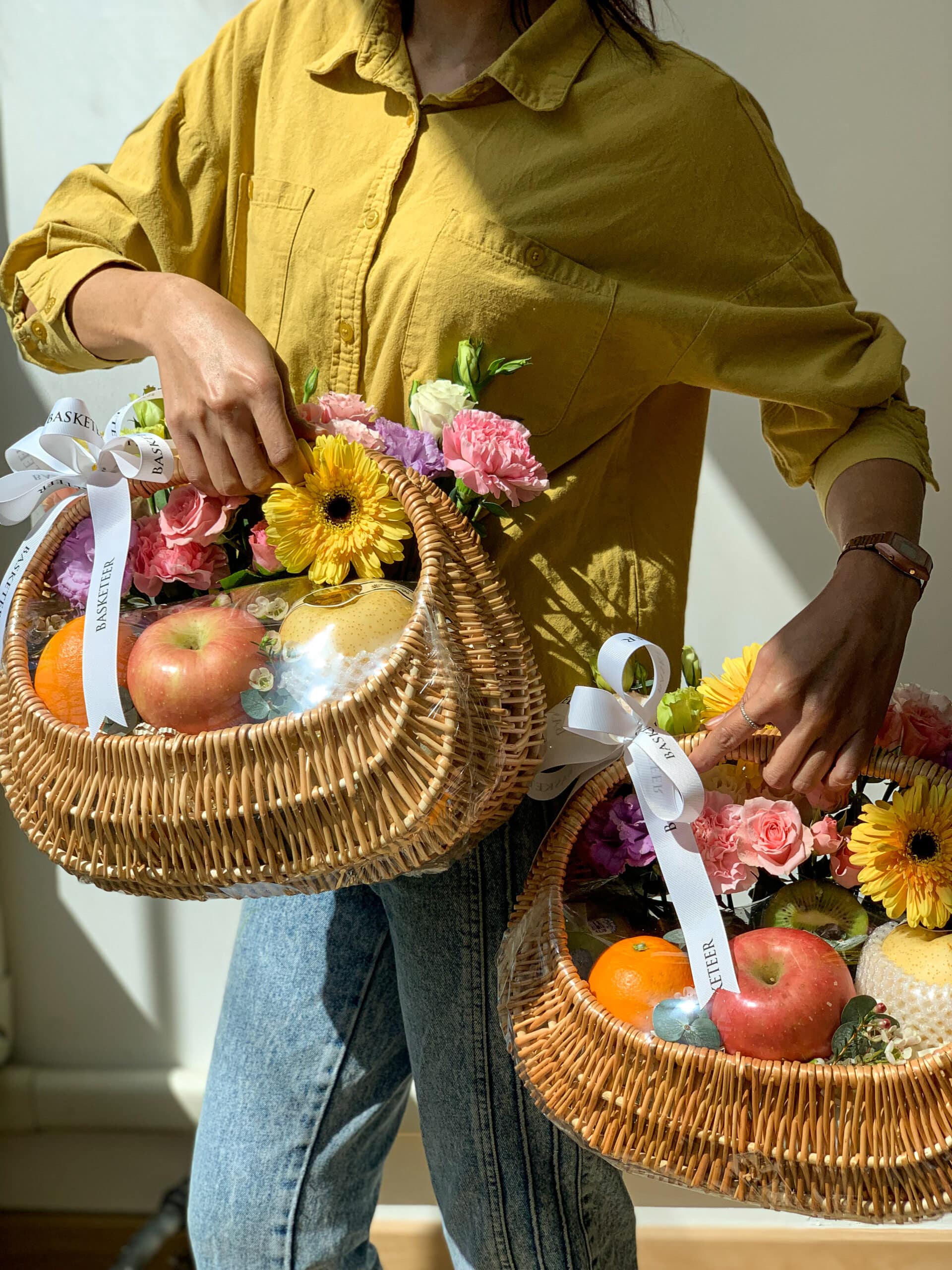 A person in a yellow shirt carries two wicker baskets filled with a luscious fruit harvest, vibrant sunflowers, and pink carnations. The baskets are adorned with white ribbons. Indoors, sunlight streams in from the side.
