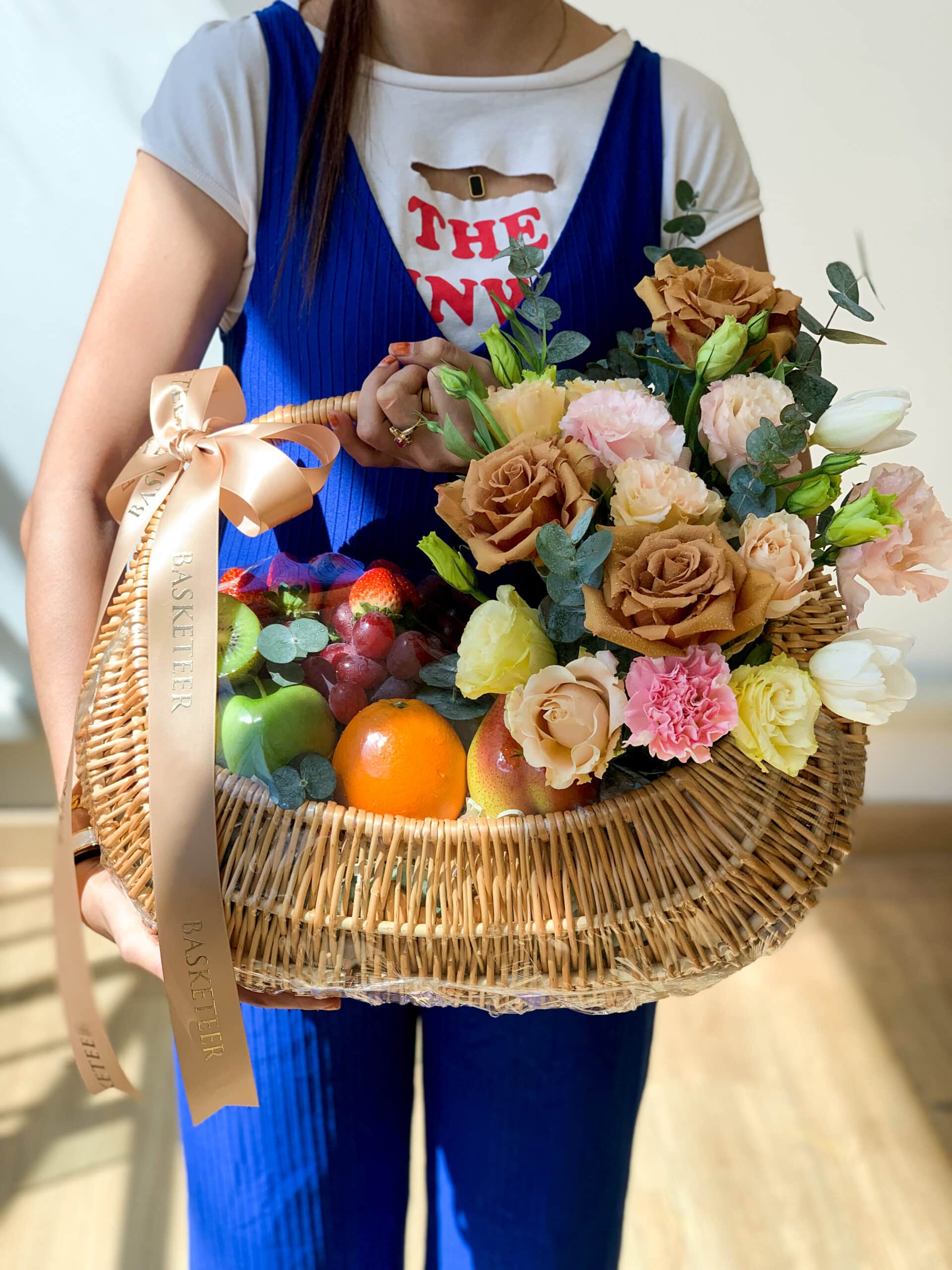 A person in a colorful outfit holds a wicker basket containing fresh fruit and a bouquet of assorted flowers. The basket handle has a ribbon with the word 