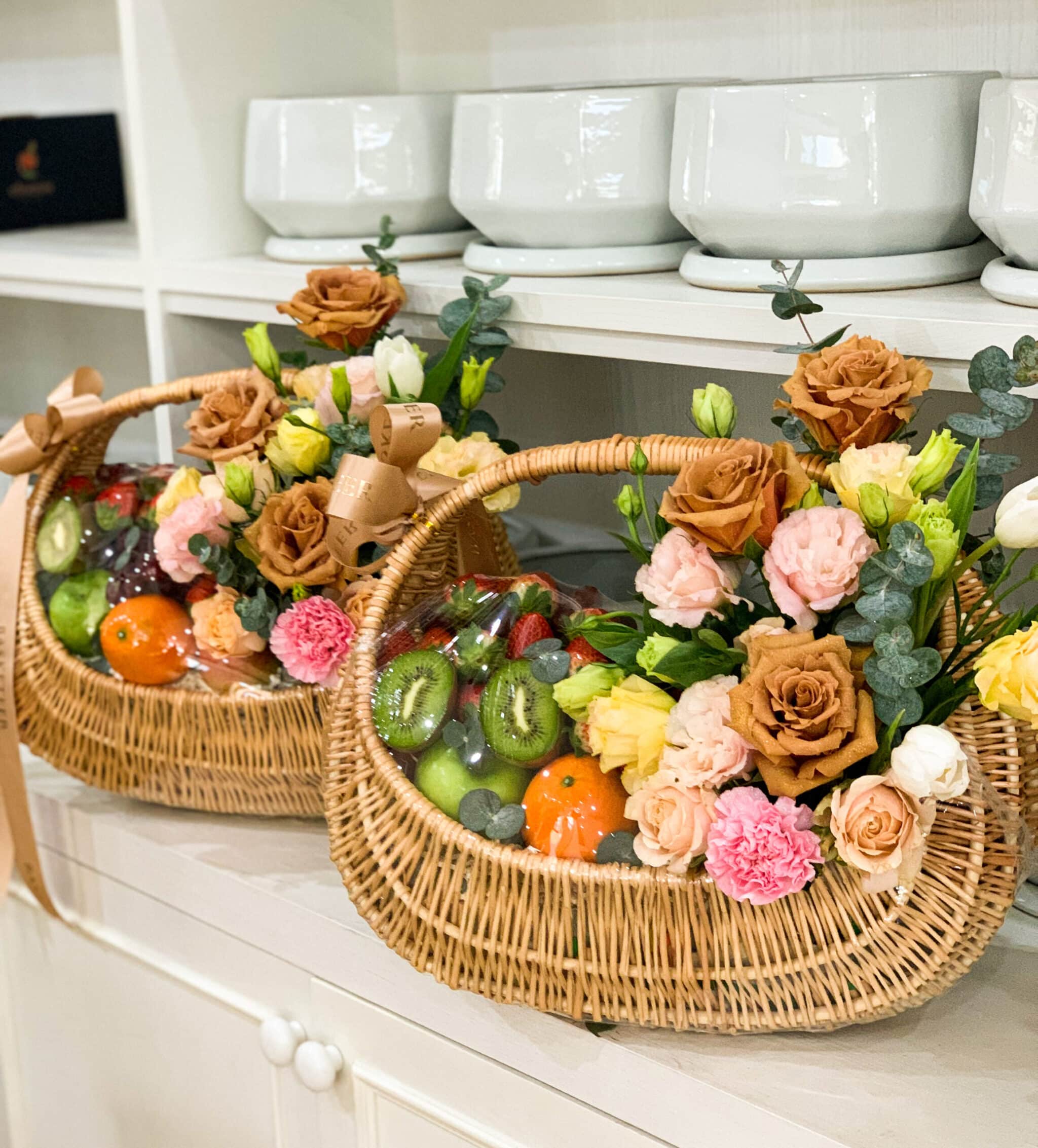 Two wicker baskets containing fresh fruit and colorful flowers are displayed on white shelves. Each basket includes items such as kiwis, oranges, and even toffees, complemented by an array of flowers like roses and carnations in shades of pink, yellow, and brown.