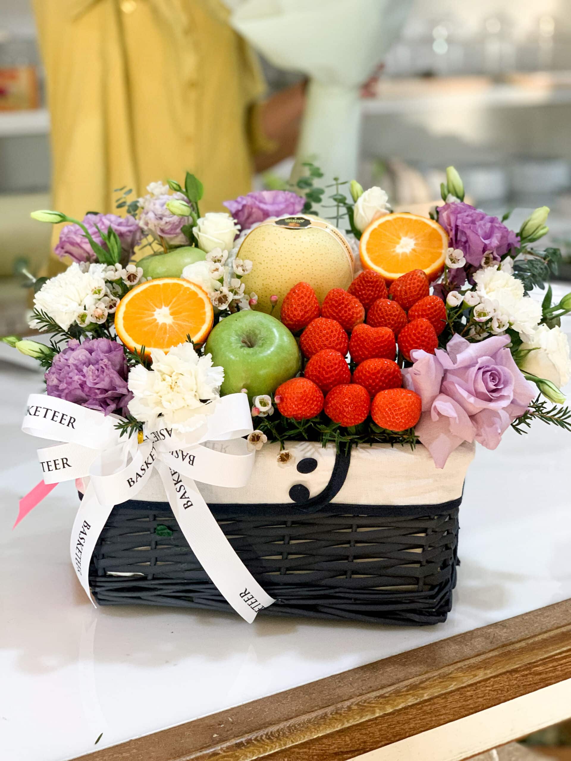 A black wicker basket filled with fresh fruits and flowers. The Purple Flowers Deluxe Fruit Basket includes green apples, a melon, orange halves, strawberries, purple and white flowers, and green foliage. The basket is adorned with a white ribbon with 