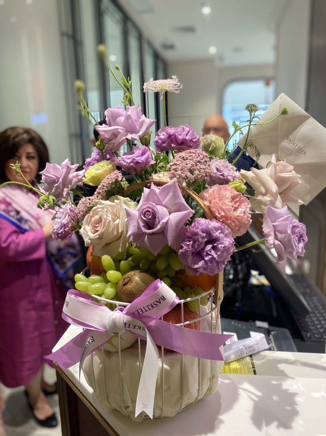 A colorful flower arrangement featuring purple blooms, pink, and white flowers sits on a counter in a basket adorned with a ribbon labeled 