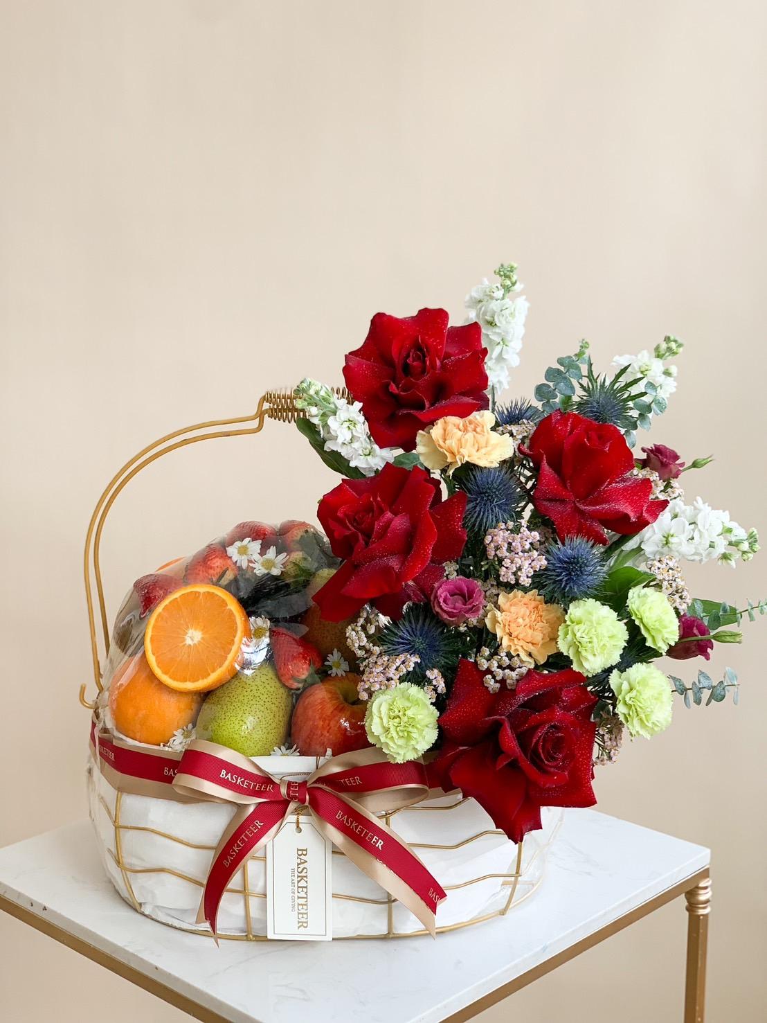 A wicker basket containing an assortment of fresh harvest fruits on the left and a vibrant floral bouquet, including red roses, white blooms, and green carnations, on the right. The basket is adorned with a red ribbon and a tag reading 