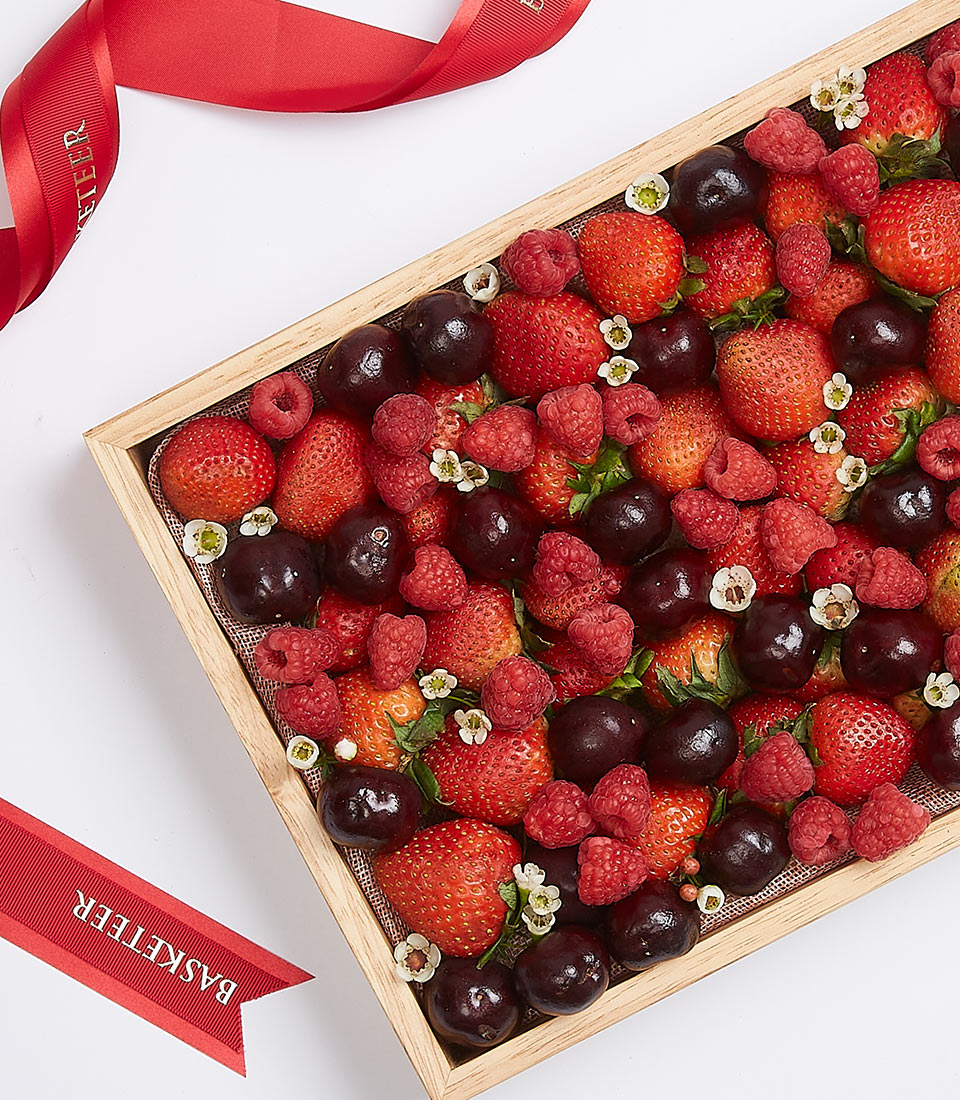 A luxurious mixed berry gift set elegantly displayed on a wooden tray brimming with luscious fresh strawberries, vibrant raspberries, and succulent dark cherries. This opulent arrangement is exquisitely garnished with delicate white blossoms. The tray is tastefully adorned with rich red ribbons accented with the distinguished 