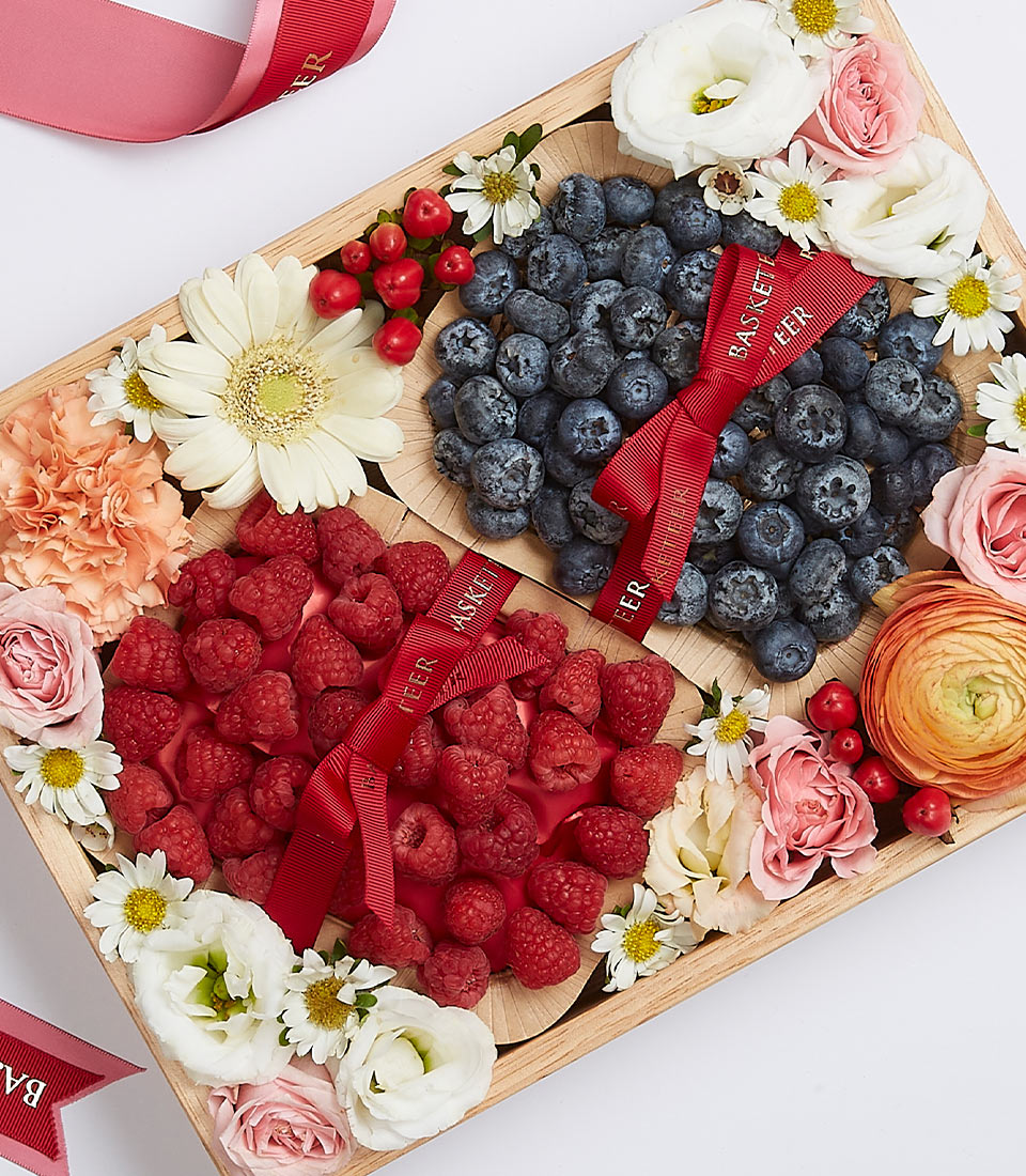 A sumptuous wooden gift box by Basketeer, elegantly designed to offer a delectable duo of fresh blueberries and raspberries. Each fruit-filled section is meticulously tied with an exquisite red ribbon, embossed with the Basketeer logo. Surrounding the vibrant berries are artfully arranged blooms—roses, daisies, and gerberas—that enhance the luxurious presentation. The entire ensemble creates an inviting scene of indulgence and elegance.