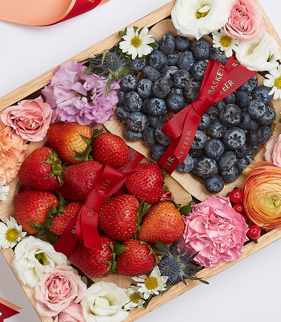 Indulge in the opulence of the Basketeer Sweet Berry Gift Set. This luxurious wooden tray, adorned with sections tied gracefully with our signature red ribbon, cradles a bounty of fresh strawberries and blueberries. Encircling the succulent fruits are an exquisite array of vibrant flowers — roses, carnations, and daisies — meticulously arranged to create a breathtakingly elegant display. An invitation to savor both beauty and flavor, perfect for any discerning palate.
