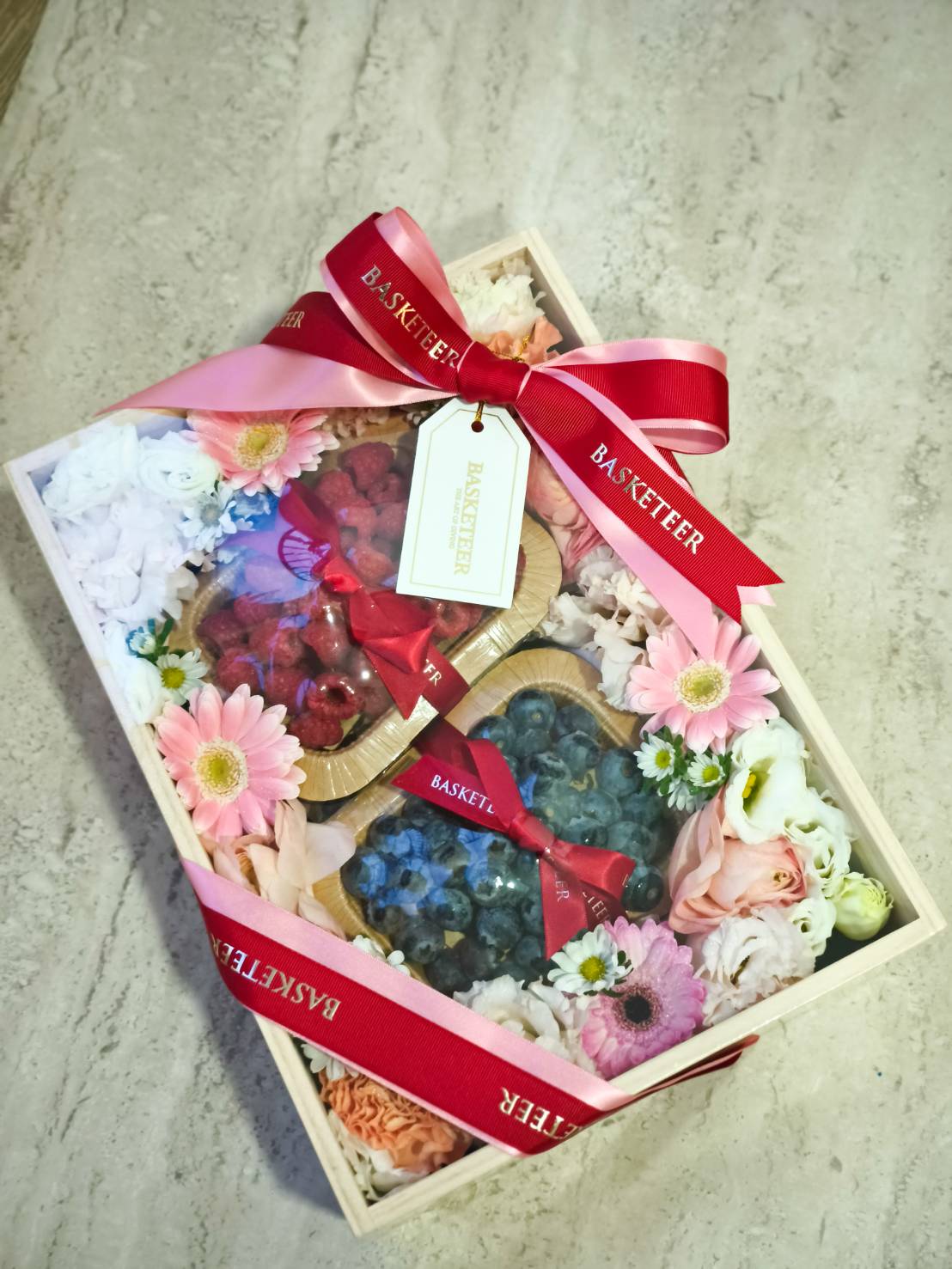 Luxury gift box with fresh raspberries, blueberries, and pastel flowers decorated with red and pink ribbons – premium fruit and flower gift