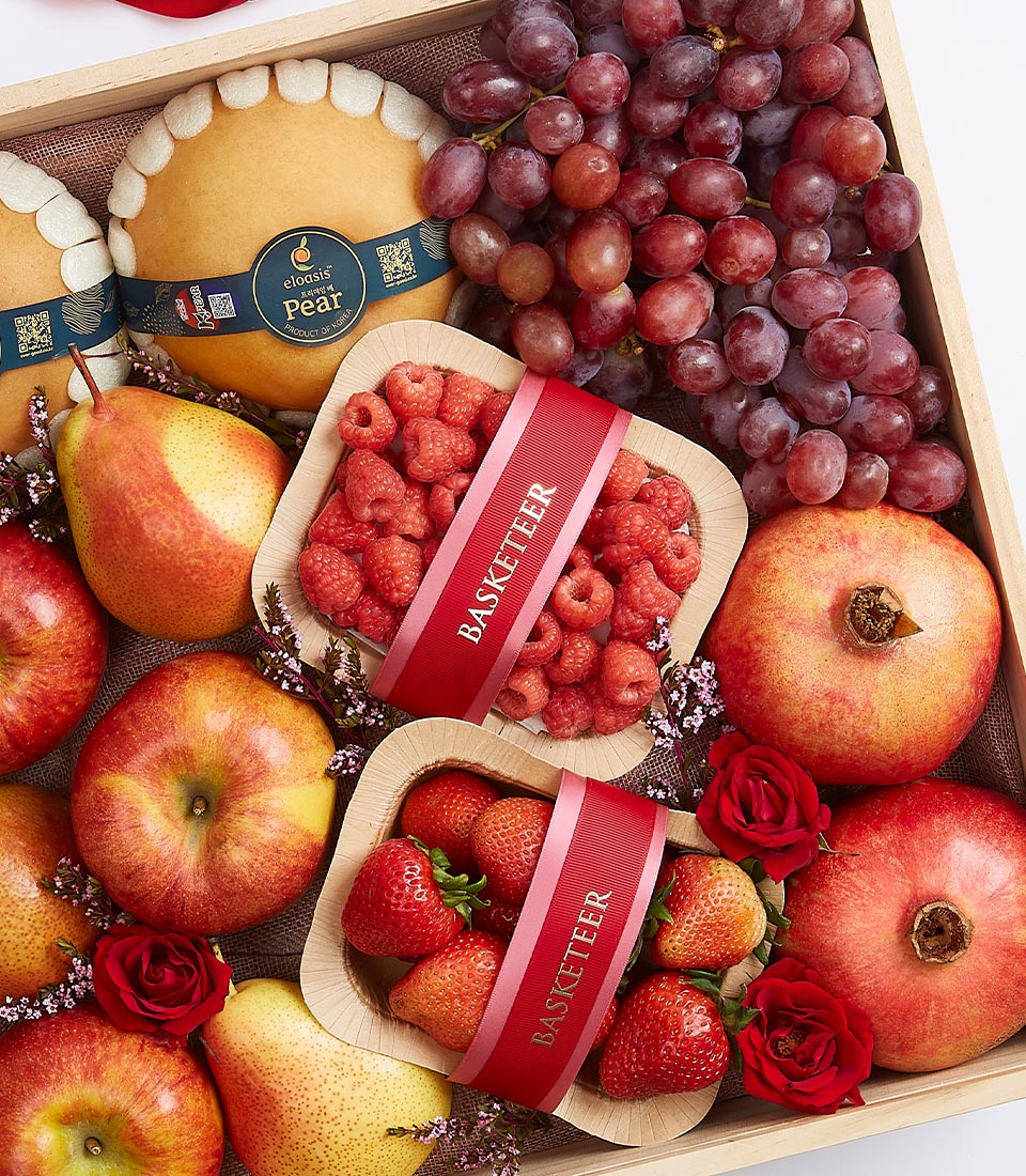 A lavish, handpicked fruit box featuring an array of premium, fresh fruits such as crisp apples, succulent pears, juicy grapes, vibrant pomegranates, plump raspberries, and luscious strawberries. These exquisite fruits are elegantly arranged around opulent red roses. The raspberries and strawberries are artfully displayed in charming wooden containers adorned with the prestigious 