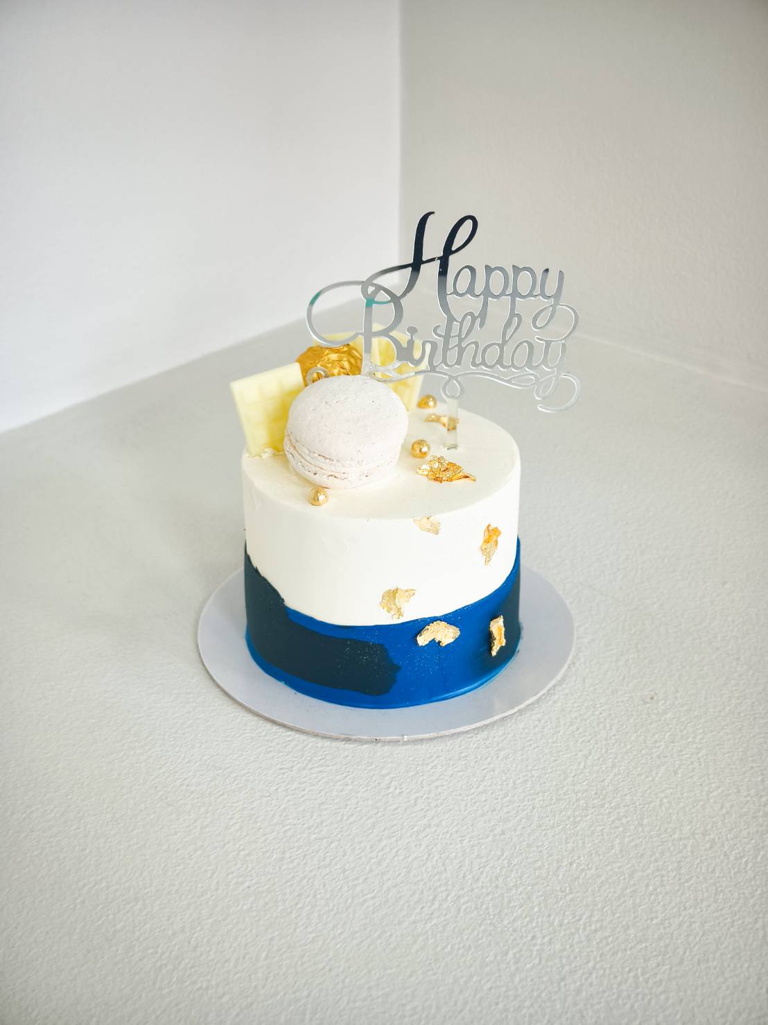 Blue and white birthday cake decorated with gold leaf, macaron, and chocolate pieces