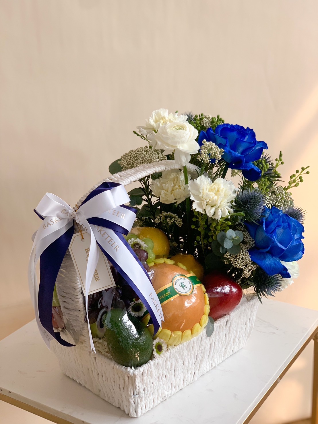 Luxury fruit and flower basket featuring premium fruits with white and blue floral arrangement, wrapped with Basketeer ribbon.