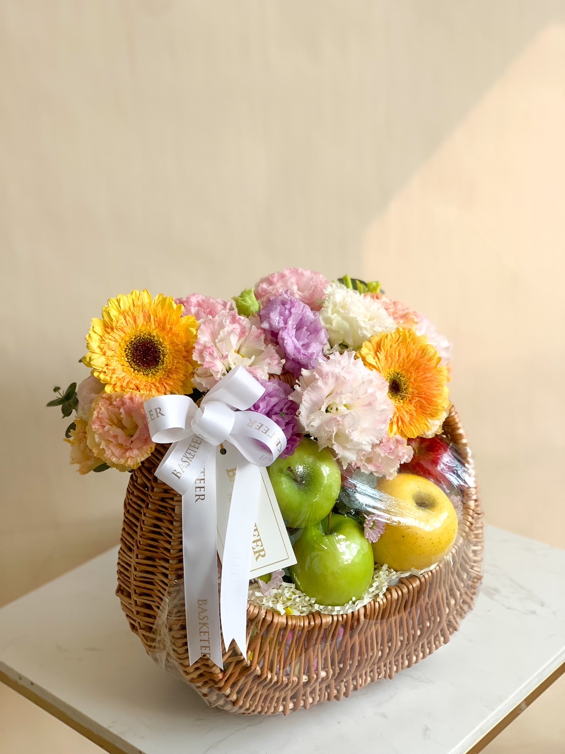 Pastel-themed fruit basket with colorful fresh flowers and assorted premium fruits in a woven wicker basket.