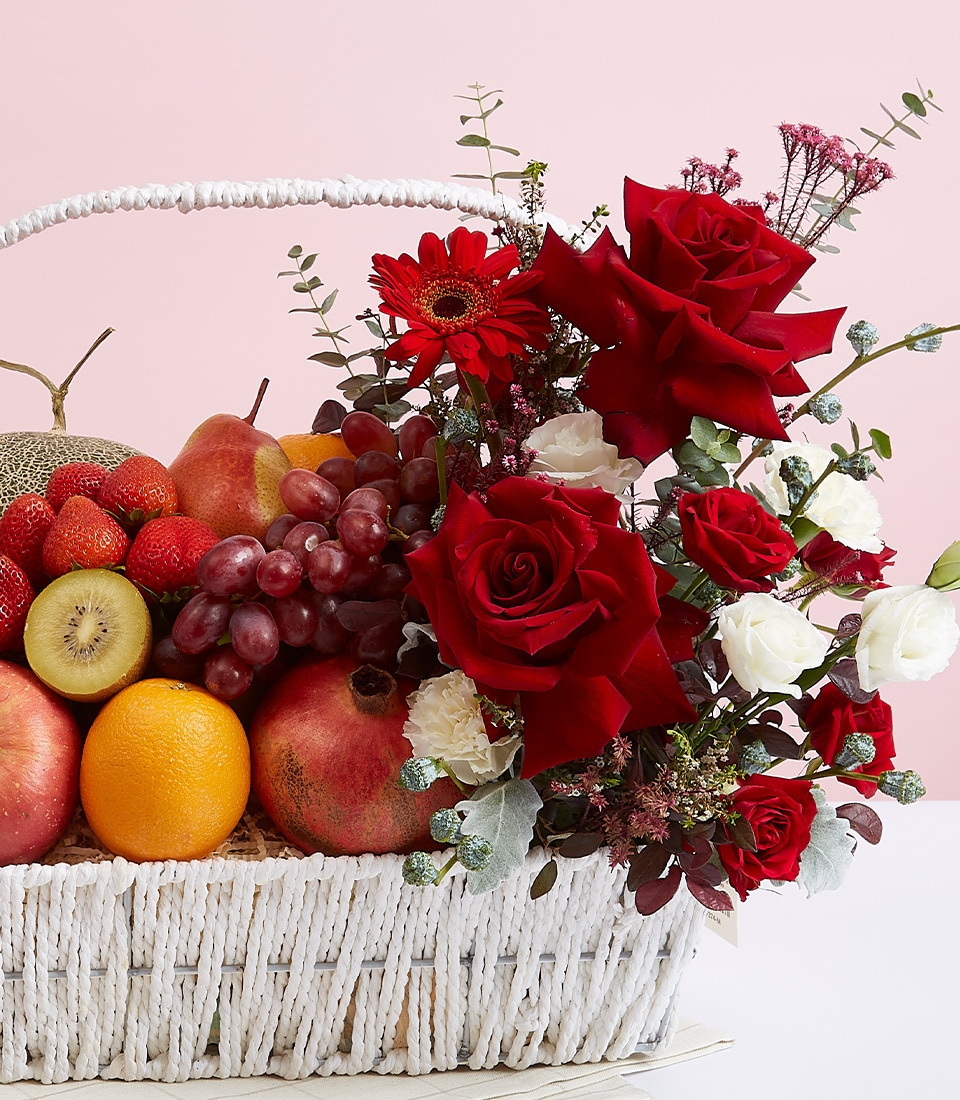 A luxurious white woven basket from Basketeer overflows with a mesmerizing assortment of the finest fresh fruits: succulent grapes, luscious strawberries, exotic kiwi, crisp apples, zesty oranges, and vibrant pomegranates. The ensemble is elevated by a stunning floral arrangement that features elegant red and white roses intertwined with lush greenery. The entire presentation is set against a soft pink background. A ribbon emblazoned with the Basketeer logo adds an extra touch of sophistication.