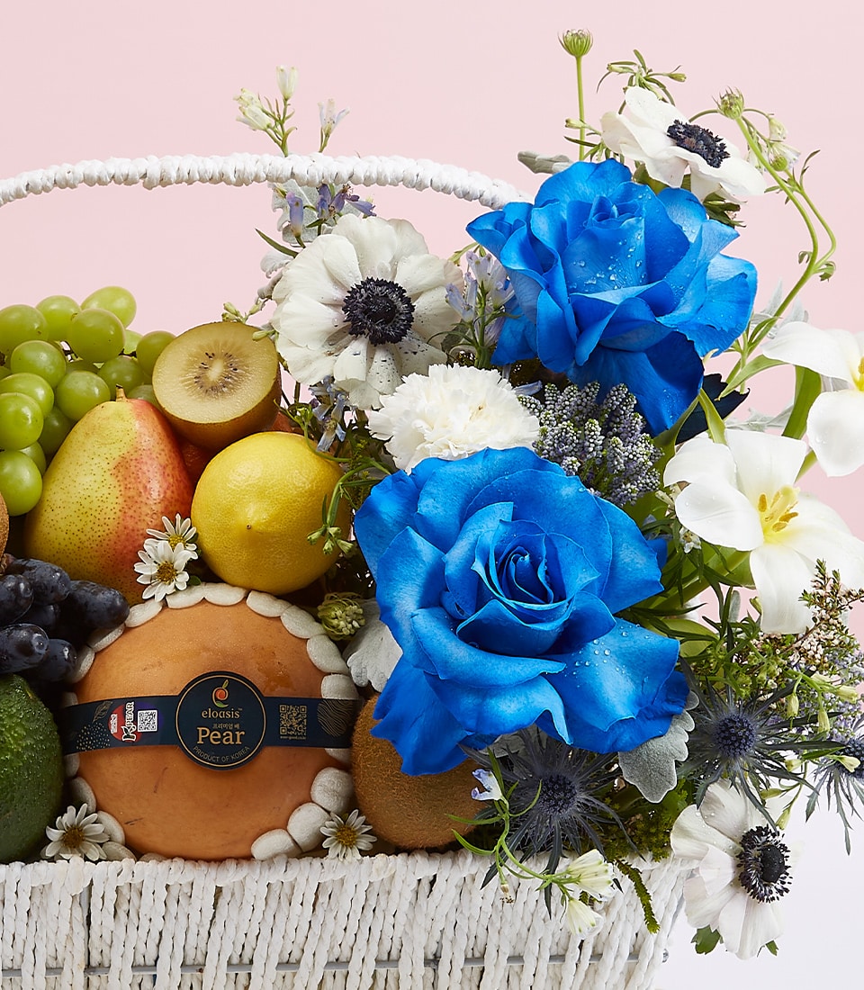 An opulent wicker basket brimming with succulent pears, luscious grapes, a crisp apple, and a vibrant orange. This exquisite Blue Rose Fruit Basket by Basketeer boasts an enchanting floral arrangement of striking blue roses and pristine white lilies, complemented by delicate blooms in a captivating display. Nestled within an elegant scene adorned with soft pink hues and the distinctive Basketeer logo ribbon gracefully tied around the box, this setting invites indulgence and elevates any occasion.