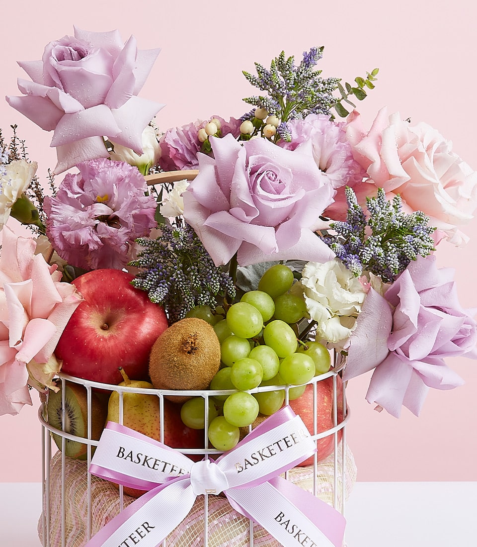 A lavish Basketeer fruit basket brimming with a delightful array of fresh apples, juicy kiwis, and succulent grapes. Elegantly adorned with light pink and lavender roses, accompanied by an exquisite selection of other flowers. The ensemble is luxuriously wrapped in a pristine white ribbon gracefully bearing the 