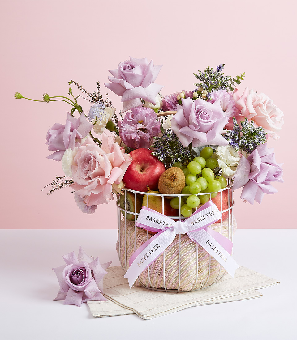 A white steel frame gift basket filled with a variety of fresh fruits such as green grapes, red apples, red pears, and many more, decorated with fresh flowers in light purple tones, with a light purple and white 