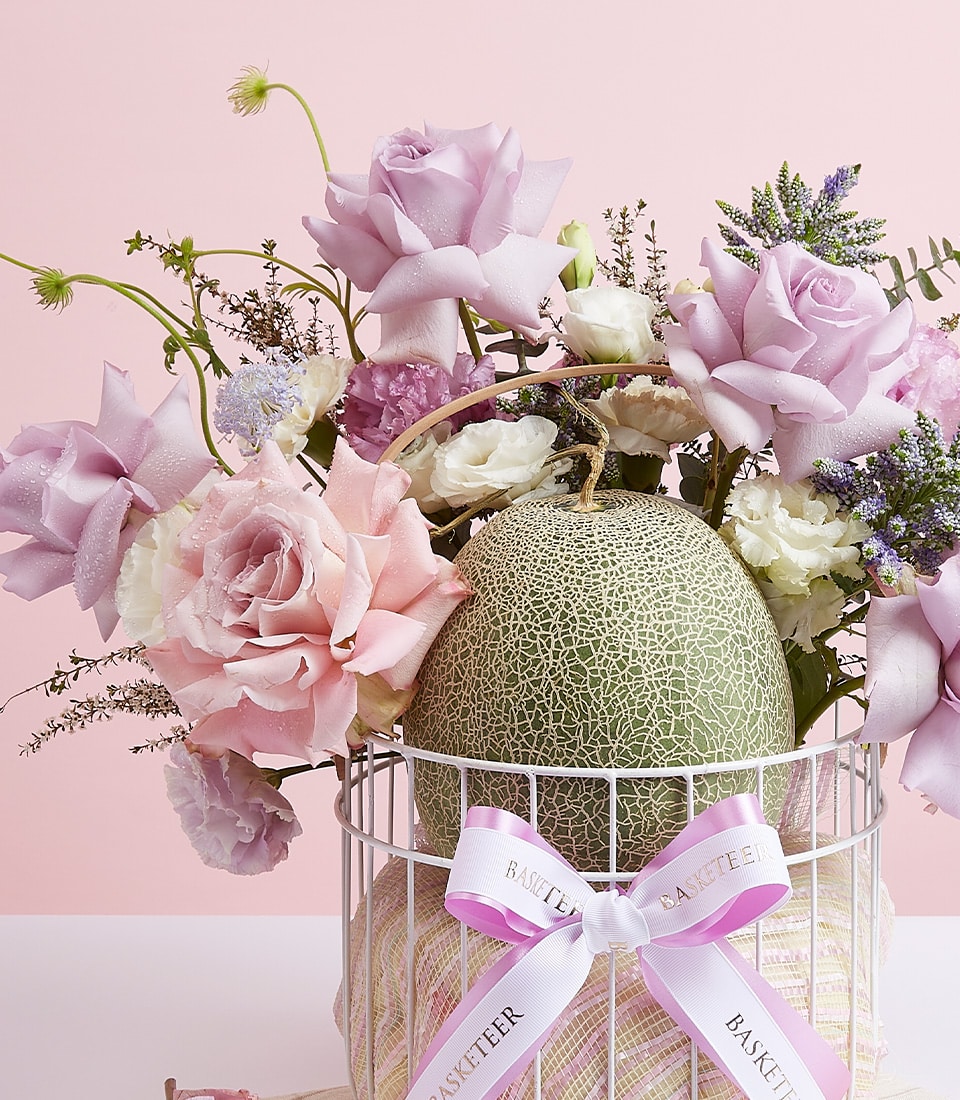 A lavish arrangement skillfully designed to captivate the senses. This exquisite Melon Fresh Fruit & Flowers Basket by Basketeer showcases opulent lavender and pink roses, ethereal white flowers, and a perfectly netted melon elegantly nestled within a pristine white lattice basket. The basket is gracefully adorned with a luxurious white ribbon bearing the distinguished Basketeer name. Set against a pastel pink backdrop, this composition exudes an inviting aura of sophistication and charm.