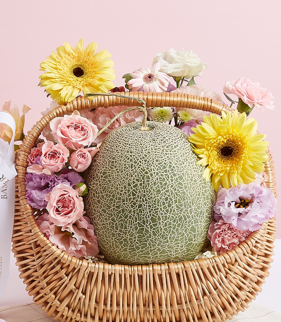 A Fresh Melon & Flower Basket by Basketeer exudes luxury and allure. Nestled within an elegantly woven wicker basket, a succulent green melon takes center stage, surrounded by an enchanting array of vibrant flowers—radiant yellow gerbera daisies, delicate pink roses, and serene pale lavender blooms. The scene is set against a soft pink backdrop that enhances the opulence of the arrangement. A sophisticated ribbon adorned with the Basketeer logo drapes gracefully across the basket, adding an extra touch of class to this exquisite ensemble.