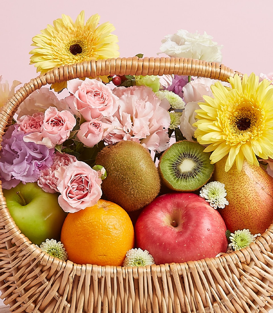 A luxurious woven basket brimming with a luscious fruit harvest features vibrant green and red apples, a zesty orange, succulent kiwis, and a perfectly ripe pear. The bountiful display is elegantly adorned with radiant yellow gerbera daisies, delicate pink roses, and soft pastel blooms. Set against an inviting light pink background, this enchanting arrangement by Basketeer exudes both freshness and charm. A tasteful ribbon bearing the prestigious Basketeer logo adds the perfect finishing touch of elegance to this exquisite presentation.
