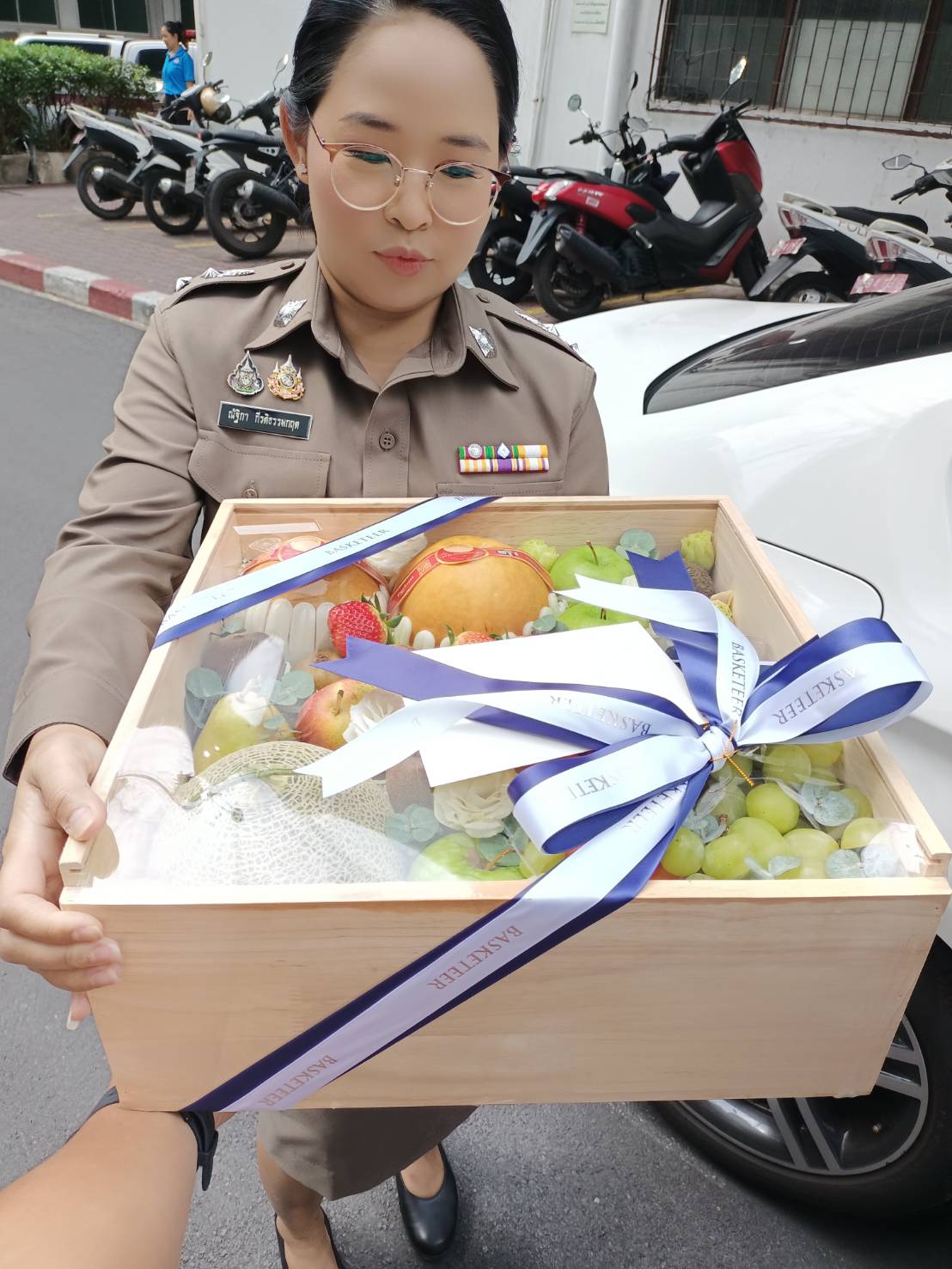 A woman in an official uniform holding a premium wooden gift box filled with fresh fruits, including grapes and a pomegranate, elegantly wrapped with a blue and white ribbon, standing outdoors near a car and motorcycles.