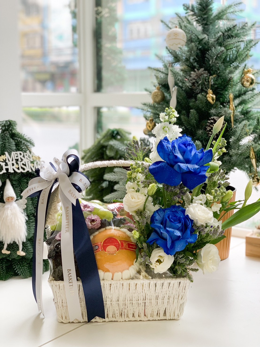 A festive gift basket from Basketeer adorned with vibrant blue and white roses, wrapped in elegant ribbons, and filled with fresh fruits and gourmet treats, set in a cozy Christmas-themed background with a decorated tree and holiday ornaments.