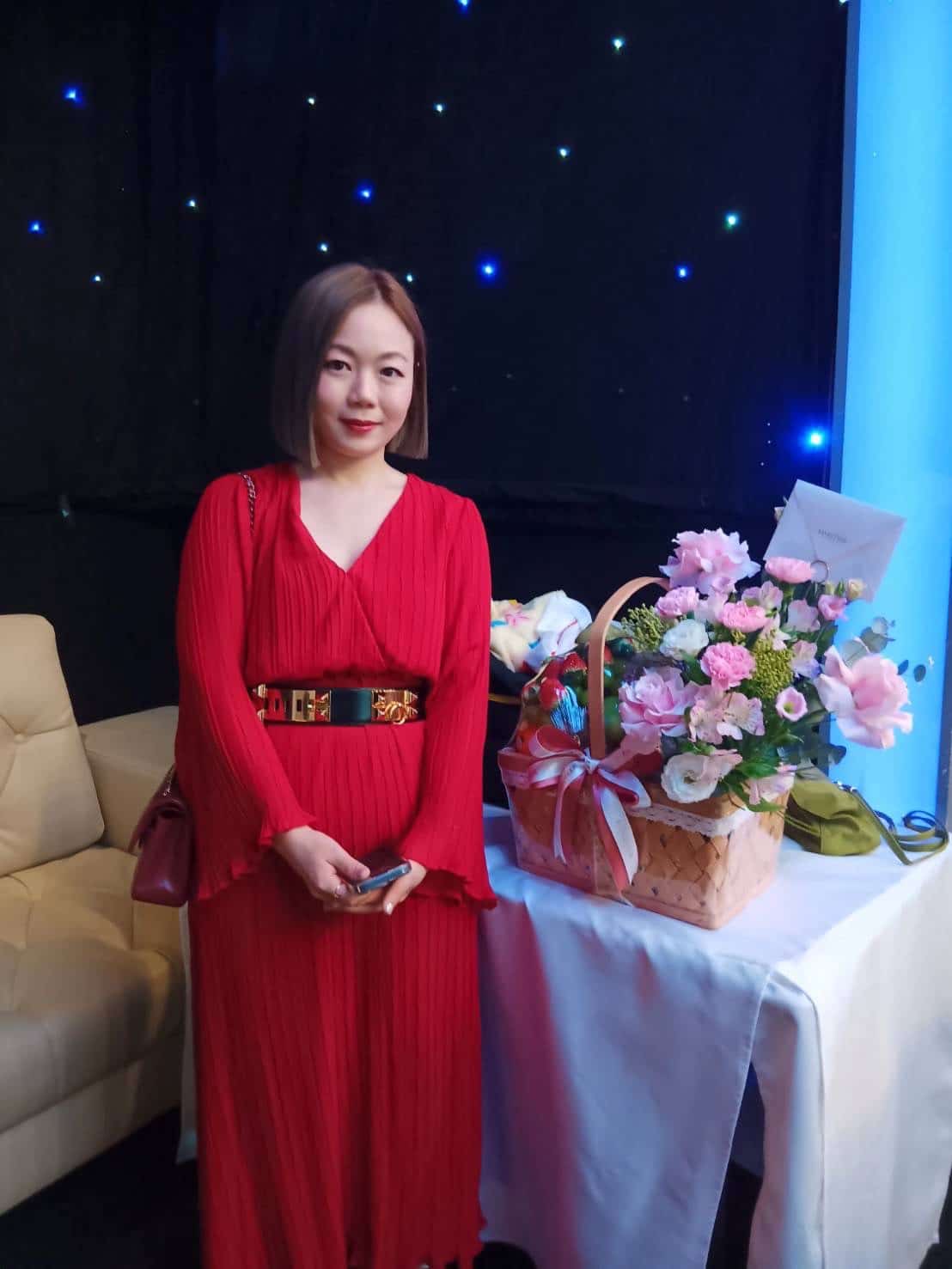 A woman in a red dress stands beside a table with a wicker basket filled with pink flowers and mixed fruits. She holds a phone in her right hand and a dark handbag at her side. The background features a black curtain with small lights and a beige couch.