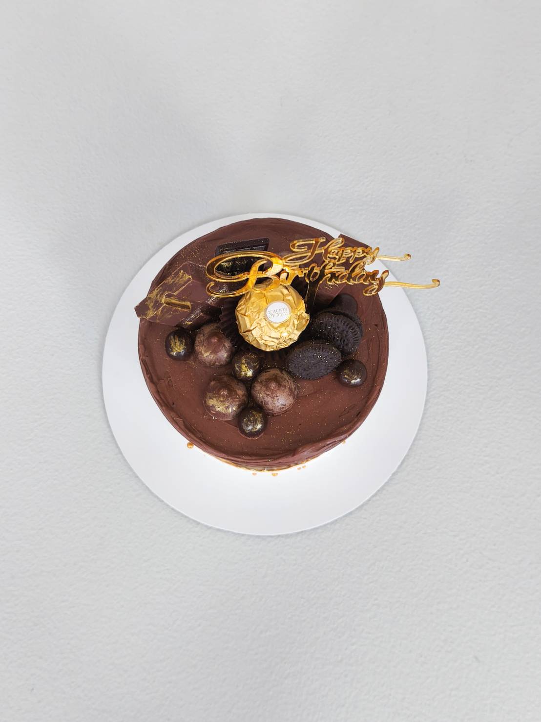 Chocolate birthday cake topped with Ferrero Rocher, Oreo cookies, chocolate balls, and golden Happy Birthday topper on white board.