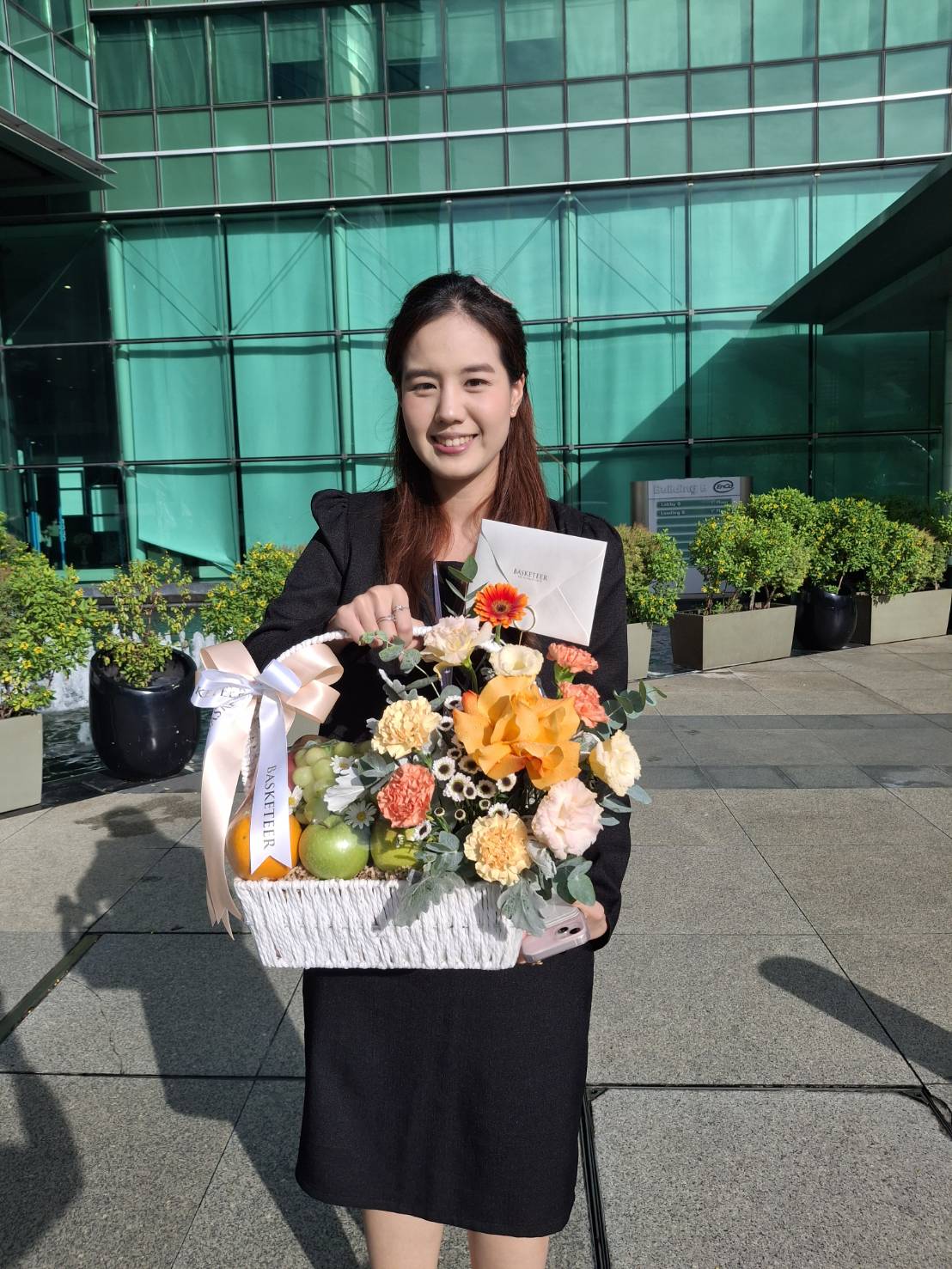 A woman in a black dress stands outdoors holding a white basket filled with colorful flowers, yellow flowers, apples, and a ribbon. She smiles at the camera, with a modern glass building in the background.