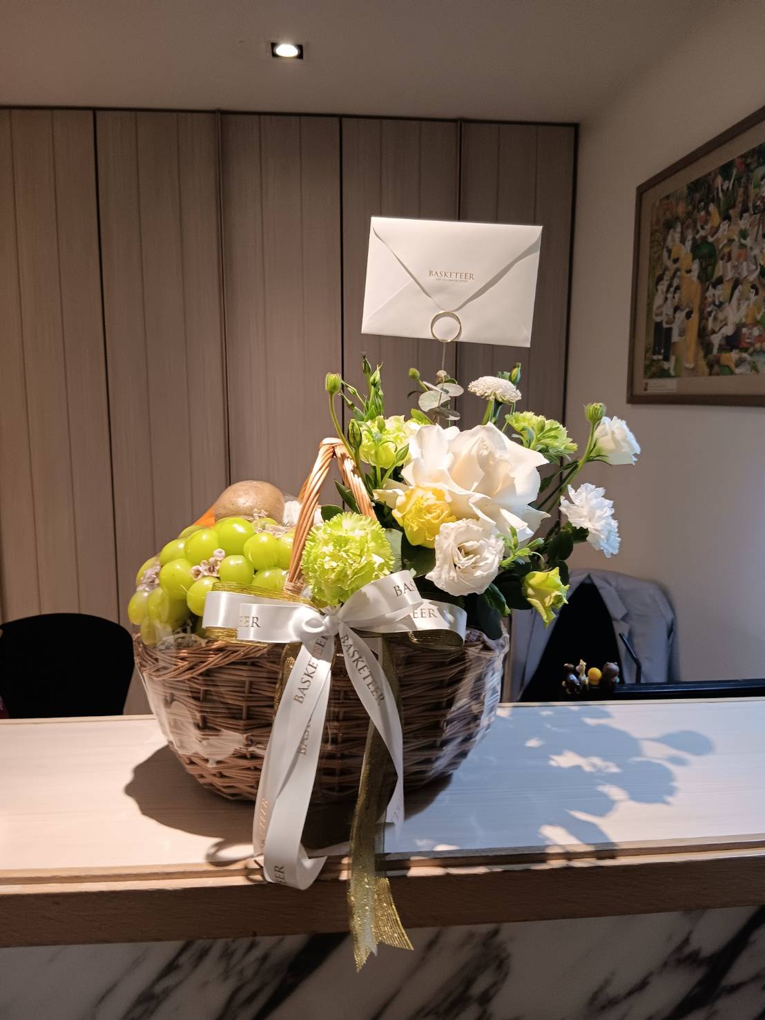 A wicker basket filled with green grapes, pears, and assorted white and light green flowers. The Fresh Fruit Rose Basket is adorned with a white ribbon and a white card labeled 