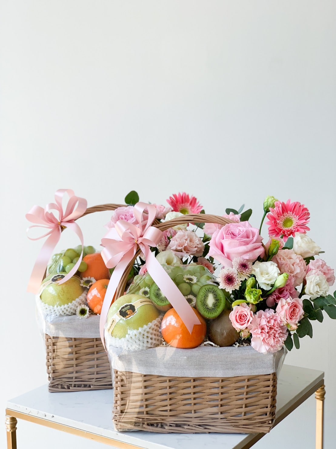 Two wicker baskets brimming with mixed fresh fruits, including oranges, kiwis, and pears, are beautifully complemented by an assortment of pink and white flowers. Adorned with pink ribbon bows, these elegant baskets rest on a light-colored surface against a plain white background.
