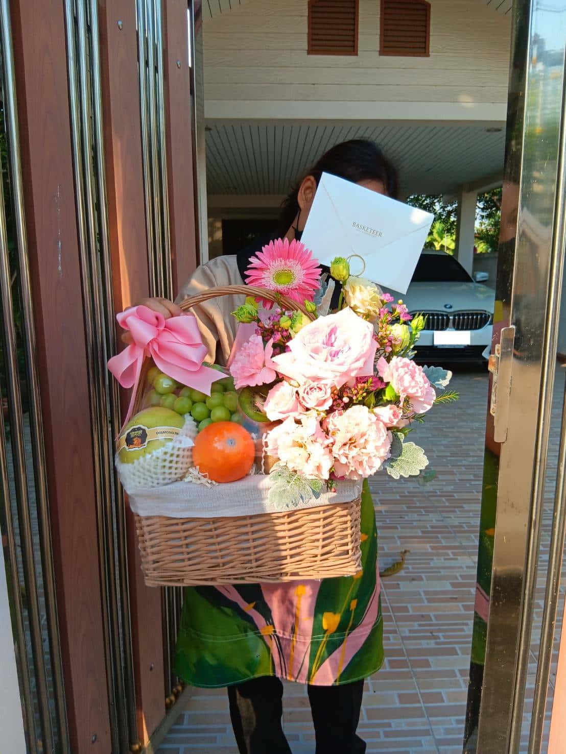 A person in colorful clothing holds a large wicker basket filled with pink roses, gerberas, green grapes, and a variety of fresh fruits. A pink ribbon adorns the Mixed Fresh Fruits Basket while the person’s face is partially covered by a white envelope bearing gold writing.