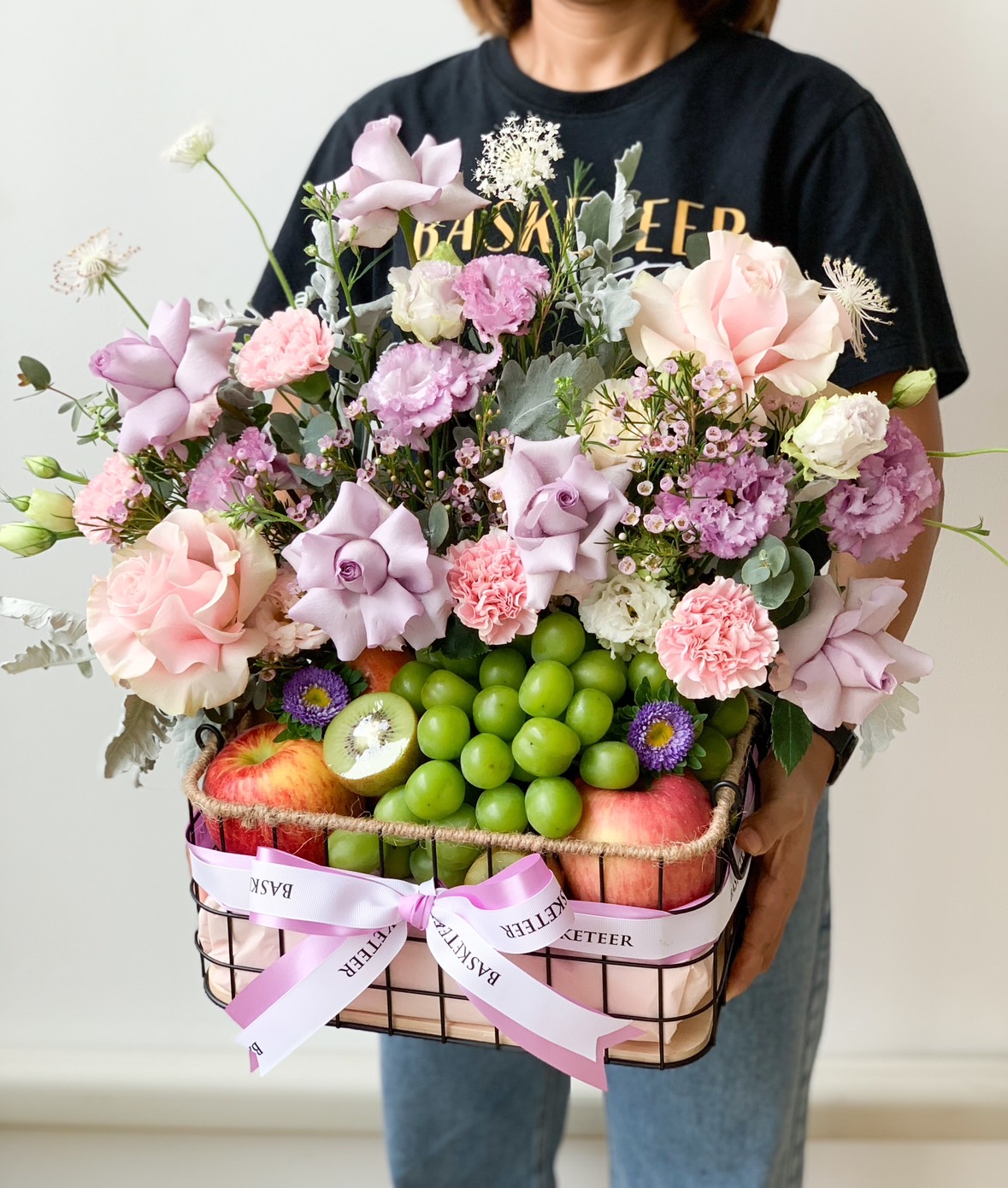 A person wearing a black t-shirt holds a wire basket filled with green grapes, red apples, and an assortment of pink, purple blooms, and white flowers. The Fruit Basket is adorned with a white ribbon that reads 