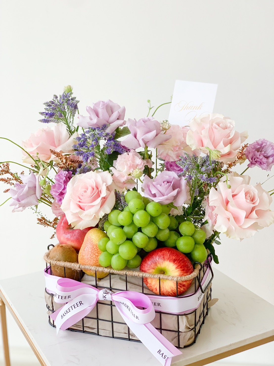 The Purple Blooms Fruit Basket features a wire container filled with an arrangement of pink roses, purple flowers, and green foliage. It includes an assortment of fruits like green grapes, red apples, and kiwis. A purple ribbon is tied around the basket, which also contains a small 