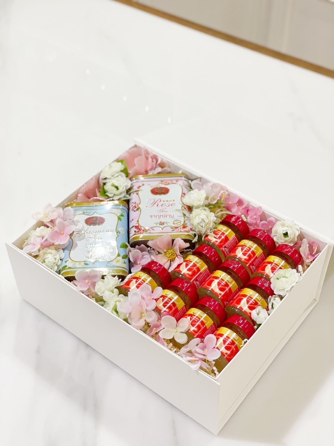 A white gift box with floral decorations containing two tins of loose tea on the left and twelve small jars with red labels on the right. The box is presented on a white marble surface.