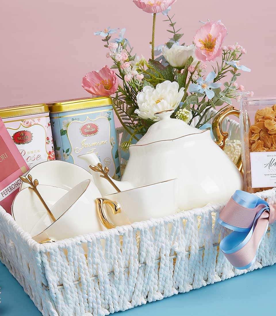 A luxurious white woven Basketeer gift basket elegantly cradles a pristine white teapot and two exquisite teacups adorned with opulent gold handles. Nestled alongside are aromatic lavender sachets, premium tins of Jasmine and Rose tea, and a charming petite bouquet of pink, white, and blue blossoms. The entire ensemble is gracefully accentuated by an enchanting pink and blue ribbon, exuding a sense of sophisticated indulgence.