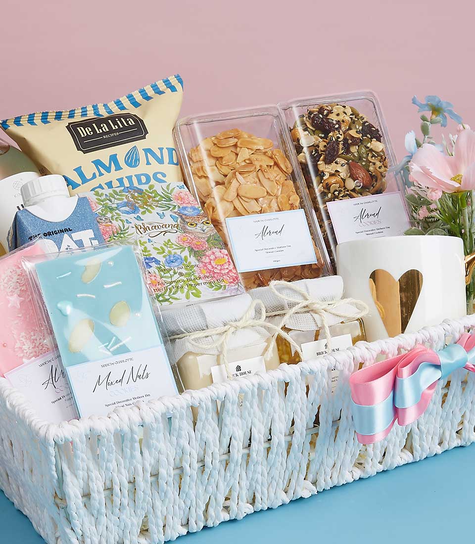 An exquisite white woven gift basket holds a curated selection of gourmet snacks and indulgent items. Delight in the savory almond chips and mixed nuts, pamper yourself with luxurious soap bars, and embrace the plush comfort of a pink towel. The basket also features an elegant decorative box adorned with a gold heart, nestled among beautiful flowers. The ensemble is beautifully finished with a sophisticated pink, blue, and white ribbon that adds a touch of grace. Set against a light pink backdrop, this lush presentation showcases the hallmark of Basketeer luxury with every detail meticulously crafted to offer an inviting experience.