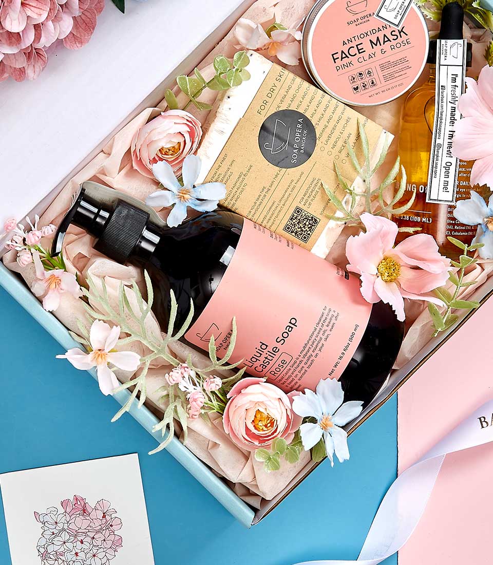 A lavish gift box exuding elegance and refinement, featuring a selection of luxurious items: a bottle of liquid hand soap, an indulgent face mask, a fragrant bar soap, and a sleek serum bottle. The pristine presentation is enhanced by the surrounding pink and blue blossoms. Nestled within crinkled tissue paper, every detail reflects careful thought from Basketeer. A beautifully illustrated note card with floral designs adds a personal touch to this exquisite collection.