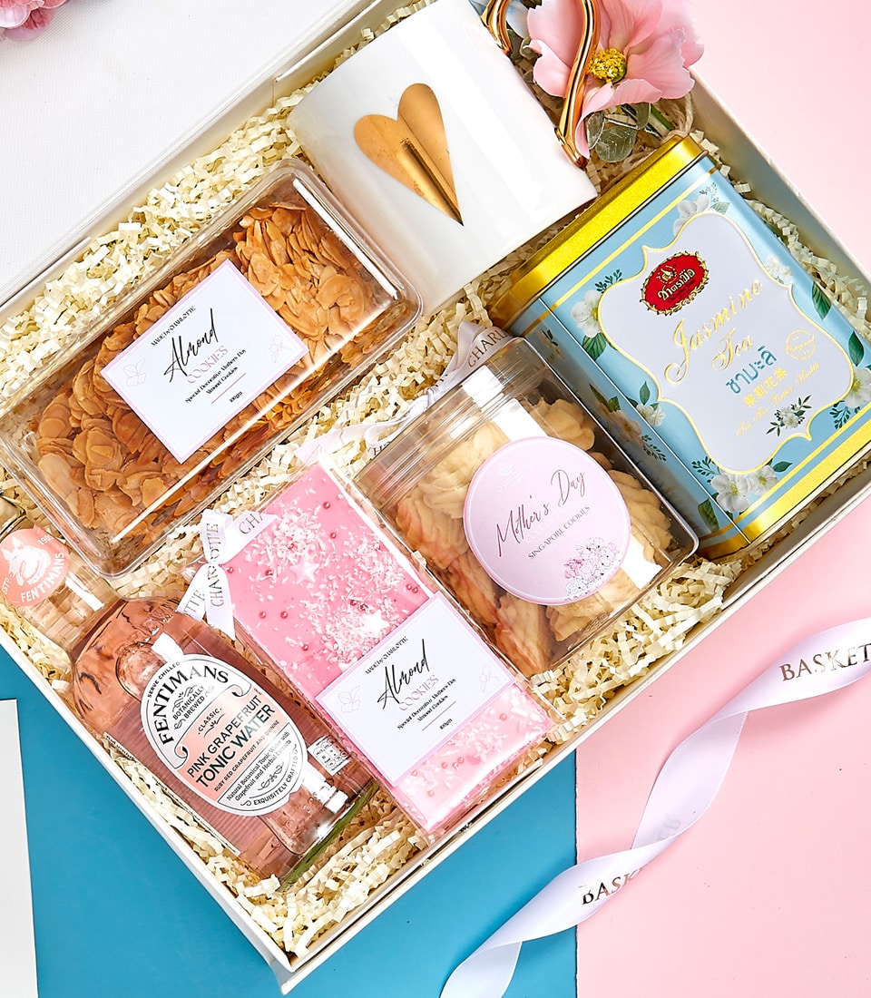 A luxurious gift box elegantly displayed on plush straw bedding, curated by Basketeer. Inside, discover a pristine white mug adorned with a delicate gold heart, an exquisite pink flower exuding grace, and a charming yellow tin with an intricate floral design. Indulge in the sophistication of a rose-flavored drink and savor the delightful almond snacks. Celebrate Mother's Day with an exclusive themed item nestled within. The presentation is completed with a beautiful pink and white ribbon bearing the distinguished Basketeer logo for that perfect touch of elegance.