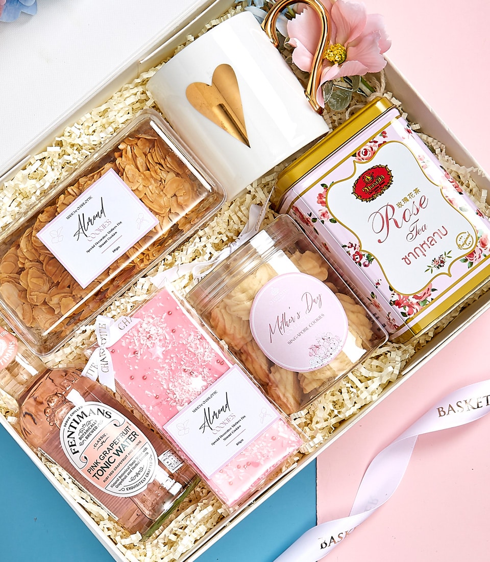 A meticulously curated gift box exudes luxury and charm, featuring a pristine white mug adorned with an elegant gold heart. Nestled within are two jars of gourmet almond snacks, alongside a beautifully packaged pink-labeled soap and a floral-scented bottle offering an indulgent sensory experience. A chic tin of rose tea adds a touch of sophistication, all complemented by a delicate ribbon inscribed with 