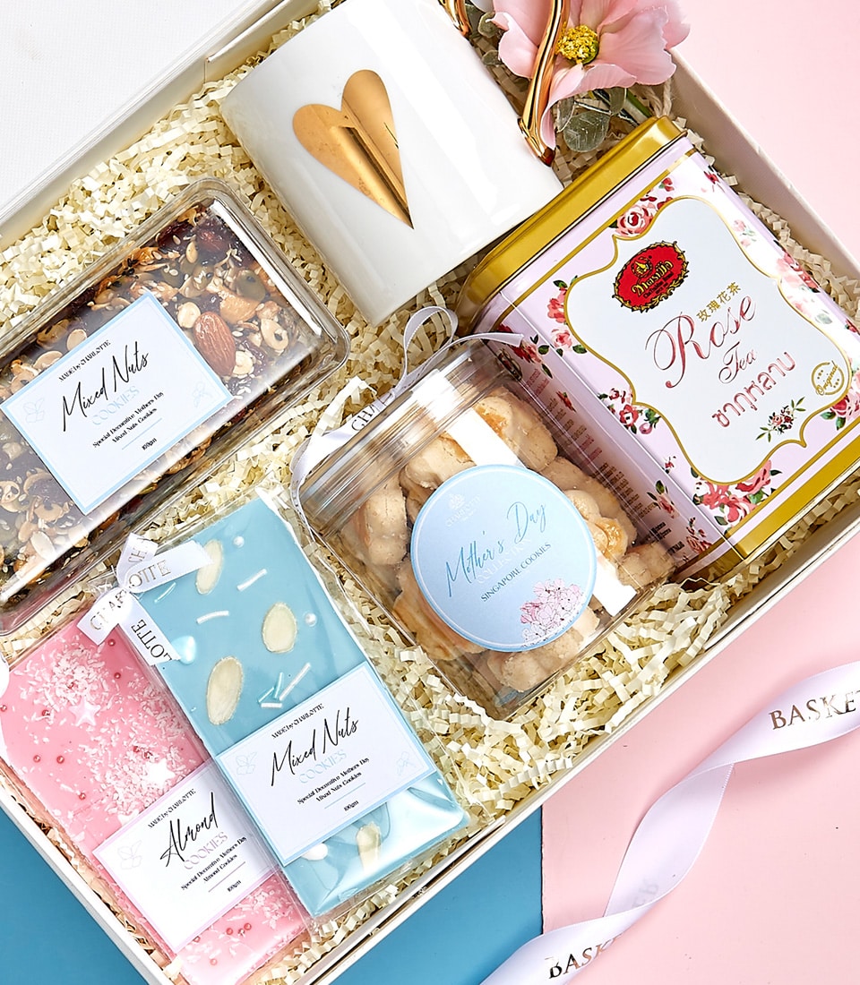 A luxurious gift box from Basketeer gracefully arranged on a bed of elegant shredded paper. Inside, you will discover a stunning assortment of delights: a pristine white mug adorned with a shimmering gold heart, an exquisite tin of rose tea, artisanal jars filled with an assortment of mixed nuts and delectable sweets, and indulgent pink and blue chocolate bars. The arrangement is perfectly complemented by a delicate flower decoration. The crowning touch is the box's lid, showcasing a stylish ribbon printed with the 