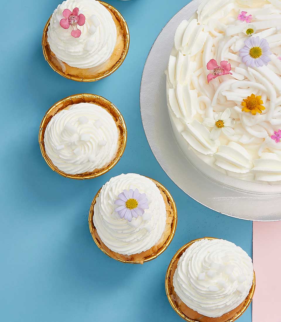 A luxurious frosted coconut cake adorned with vibrant floral decorations takes center stage on an elegantly set table, framed by a soft blue and pink backdrop. Accompanying this opulent centerpiece are four smaller coconut cakes, each exquisitely topped with white frosting and crowned with a delicate flower. Nestled among the cakes, a ribbon featuring the Basketeer logo tastefully enhances the setting, emphasizing quality and sophistication.