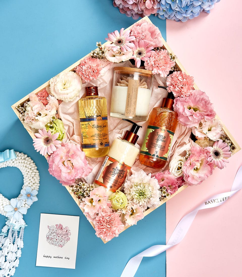Alt text: An opulent gift box adorned with the prestigious Basketeer logo, brimming with delicate pink and white flowers. Luxurious white candles and an array of indulgent bath products, including lotions and oils elegantly encased in transparent bottles with glistening golden caps, exude sophistication. A beautifully illustrated card with 