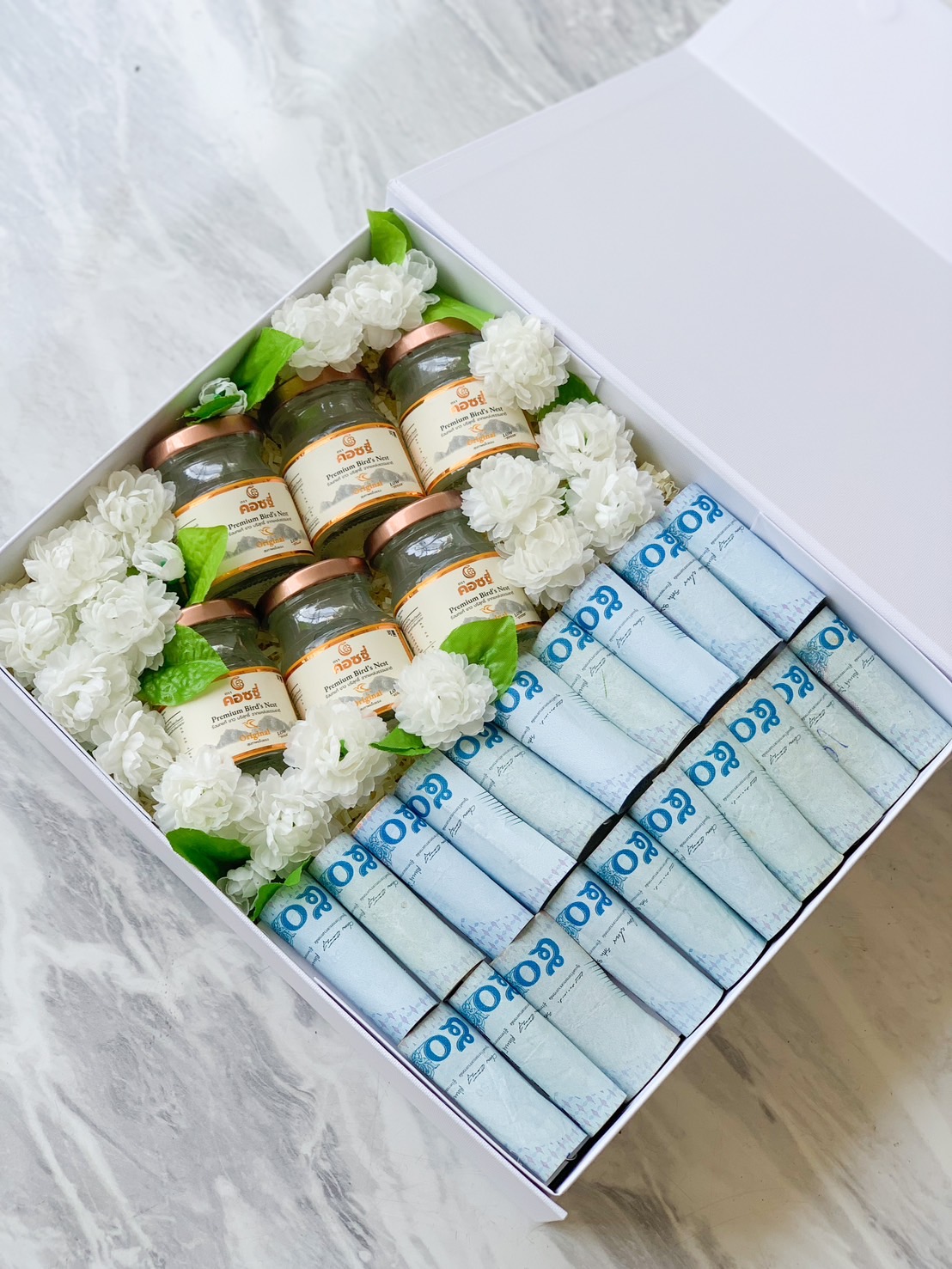 A neatly arranged gift box containing four jars of bird's nest drink, each topped with green lids and surrounded by white flowers. Beside the jars are three stacks of rectangular boxes labeled “Gao” in blue text on a light blue background. The finest gift for Mother's Day.